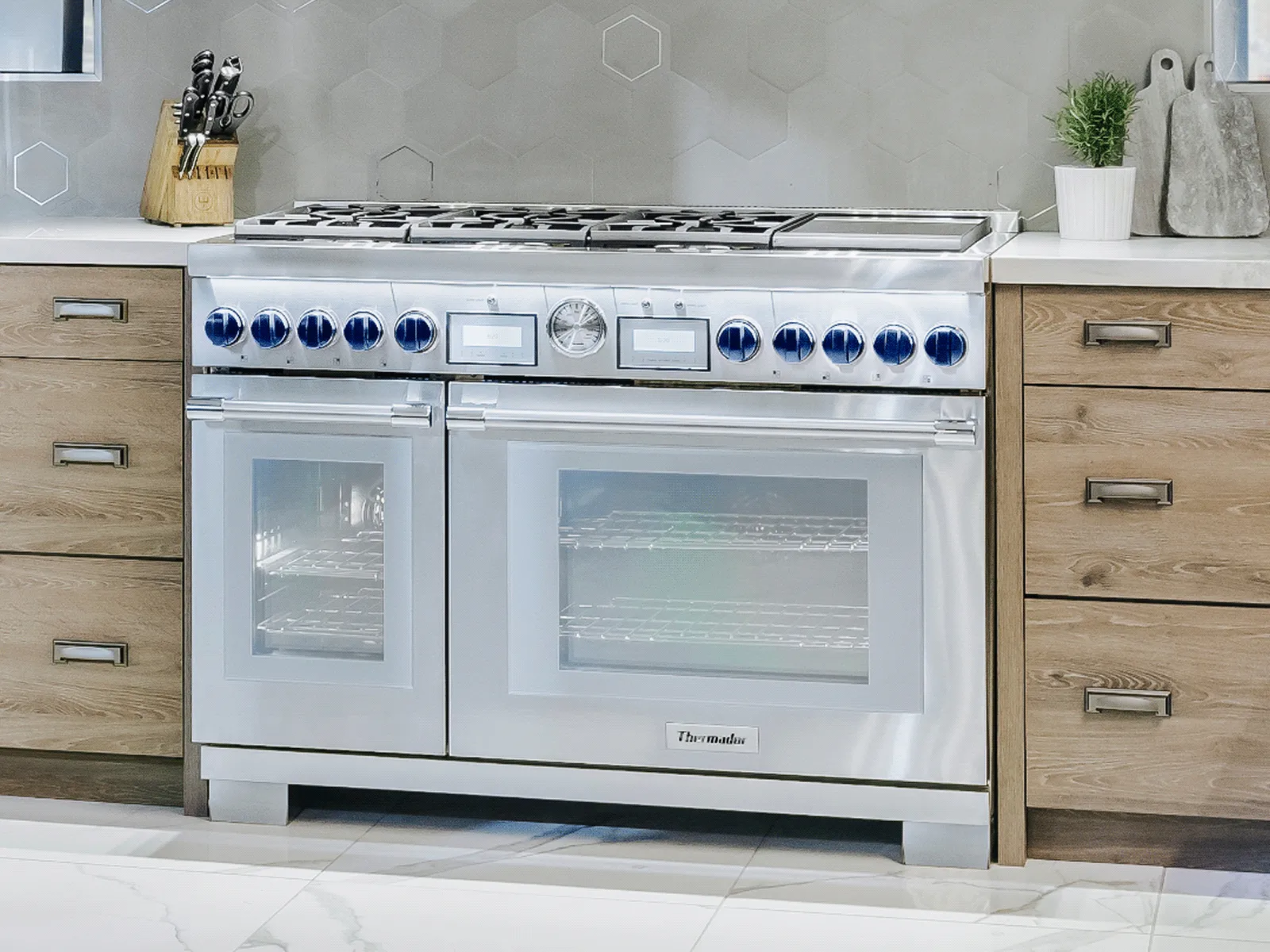 Modern stainless steel double oven range with blue knobs in a bright kitchen featuring wood cabinets and white tile flooring.