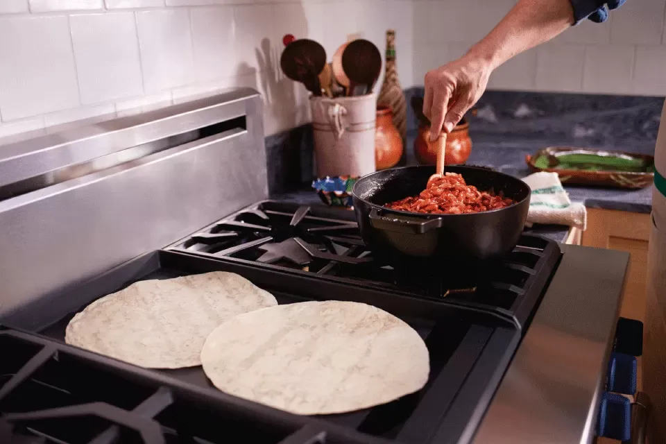Person stirring a pot of tomato sauce on a stovetop with two flour tortillas warming beside it.