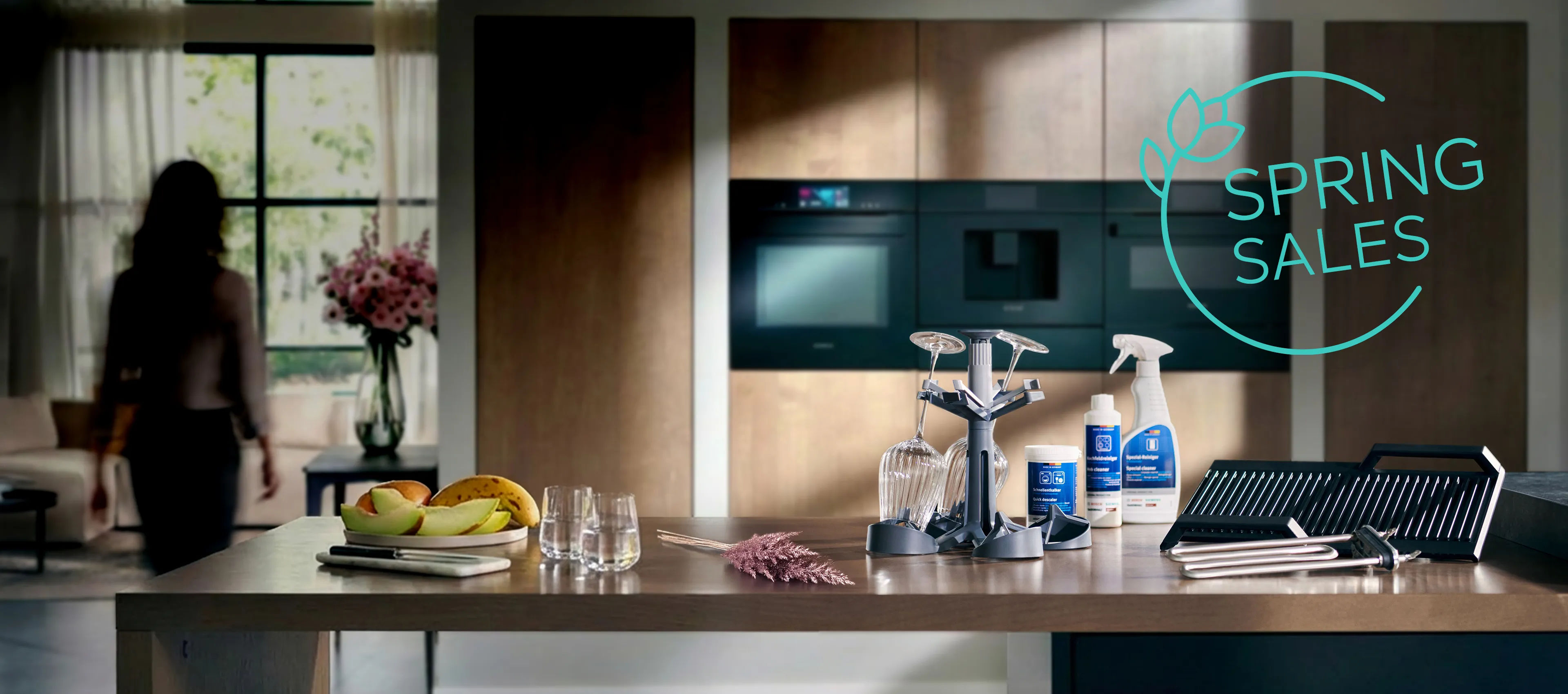 Modern kitchen countertop with cleaning products, dish rack, fruit bowl, glasses, and a woman walking near a window; "Spring Sales" text overlay.
