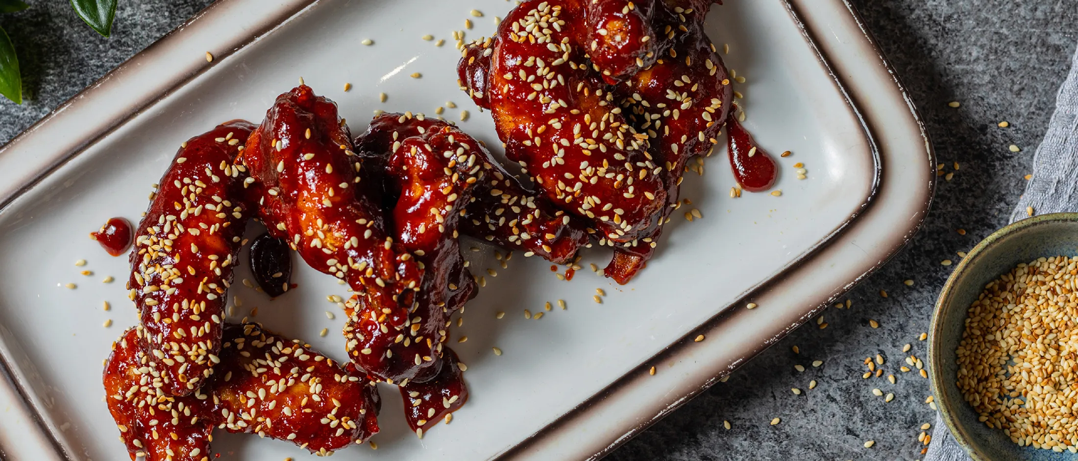 Sticky red-glazed chicken wings sprinkled with sesame seeds on a white rectangular plate, bowl of sesame seeds at the right.