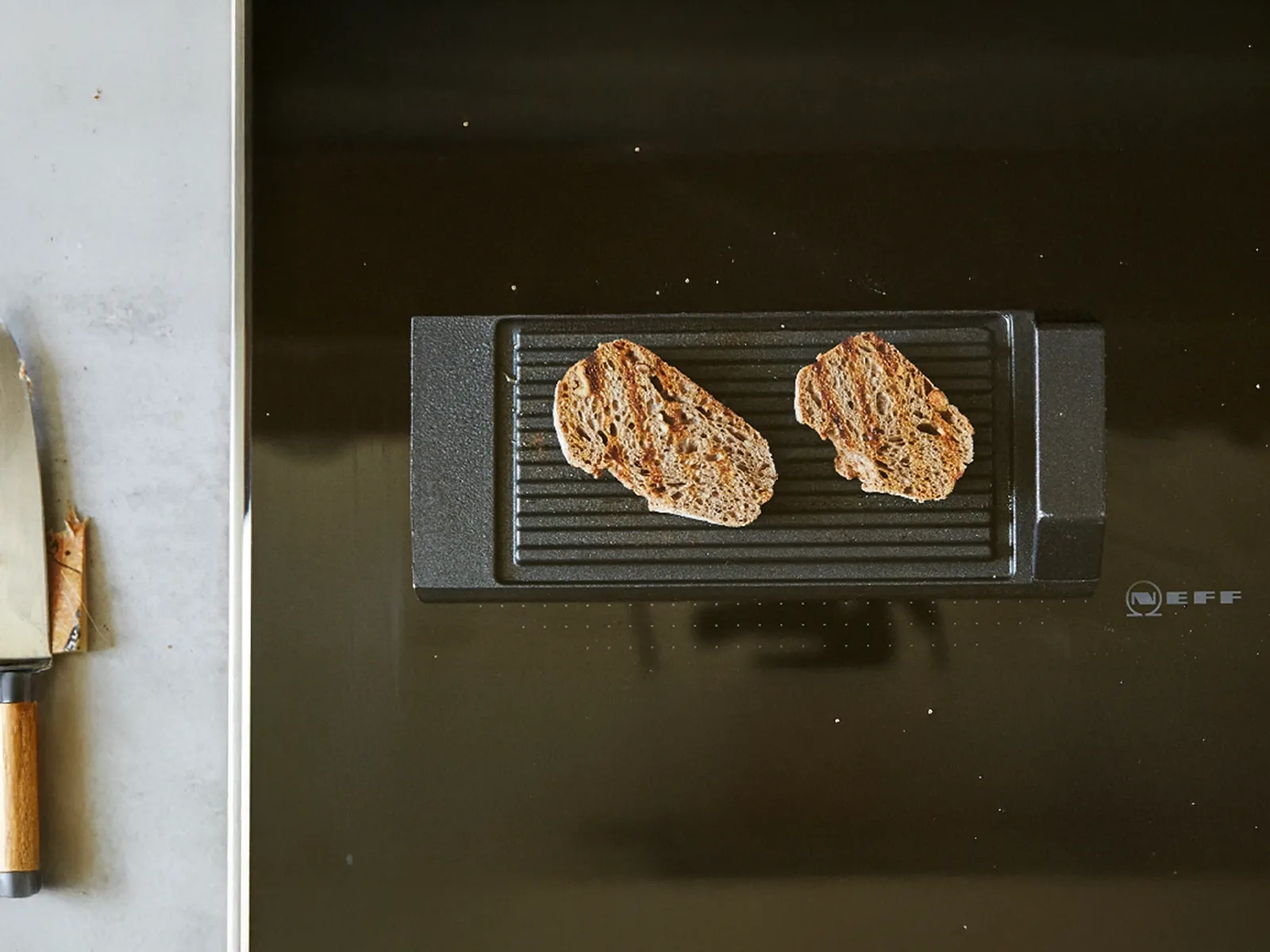 Due fette di pane grigliato su una padella grill nera da fornello, accanto a un coltello da cucina con manico in legno su un piano di lavoro grigio
