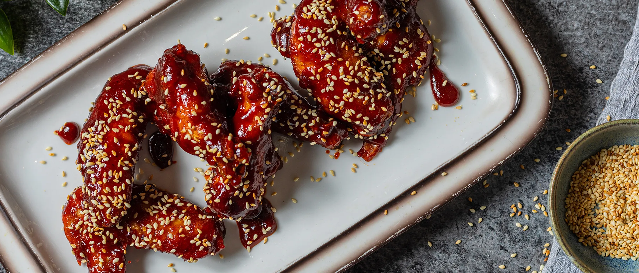 Tre pezzi di pollo glassati con salsa rossa e semi di sesamo, disposti su un vassoio rettangolare bianco; a lato una ciotolina con semi e semi sparsi sulla superficie scura.