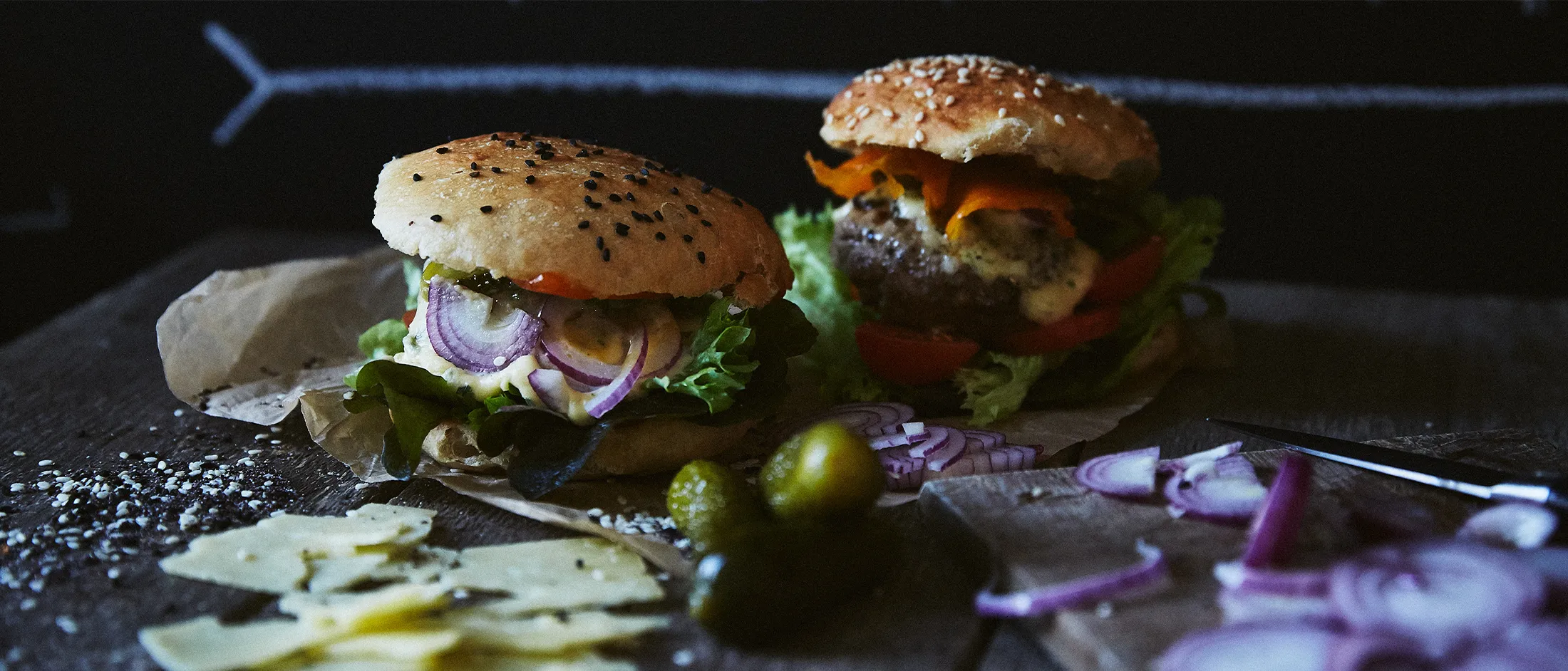 Due hamburger con lattuga, fette di cipolla e formaggio su una superficie scura con cetriolini e fette di cipolla intorno
