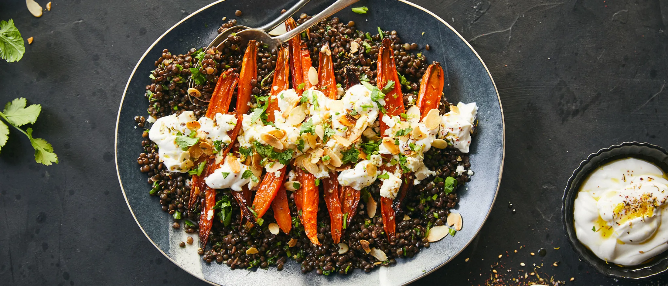 Carote arrosto, quinoa e condimento cremoso su un piatto scuro con spezie ed erbe.