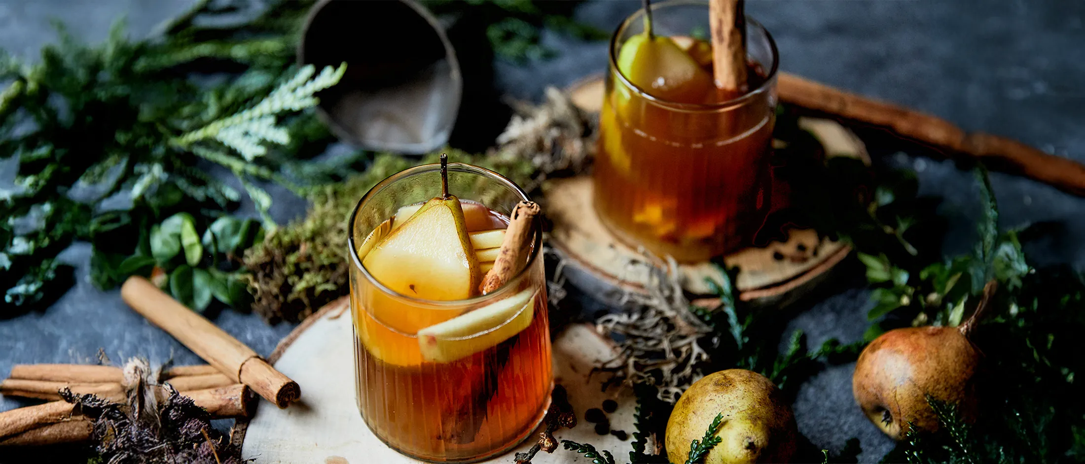 Due bicchieri con bevanda speziata con cannella e fette di pera su fette di legno circondati da frutta e spezie