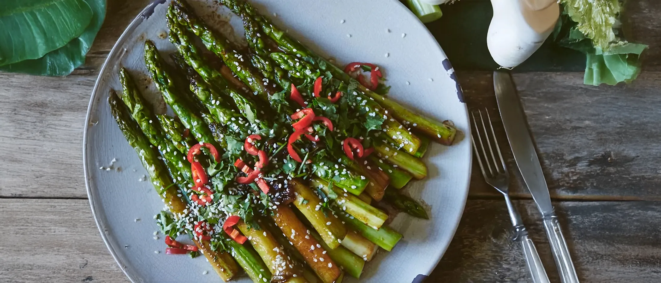 Asparagi saltati con semi di sesamo e fettine di peperoncino rosso su piatto bianco con coltello e forchetta accanto.