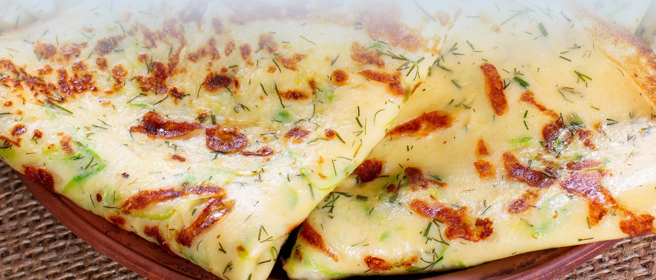 Nahaufnahme von zwei gefalteten herzhaften Crêpes mit Kräutern und gebräunten Stellen auf einem braunen Keramikteller, der auf einer gewebten Unterlage steht.
