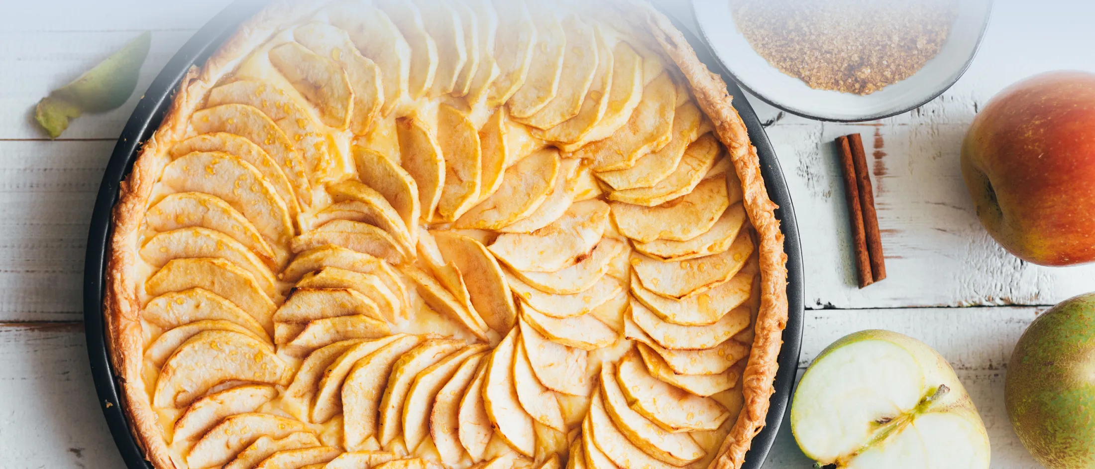 Eine hausgemachte Apfeltorte mit dünn geschnittenen Äpfeln, die spiralförmig auf einem Holztisch angerichtet sind, daneben liegen frische Äpfel und Zimtstangen.