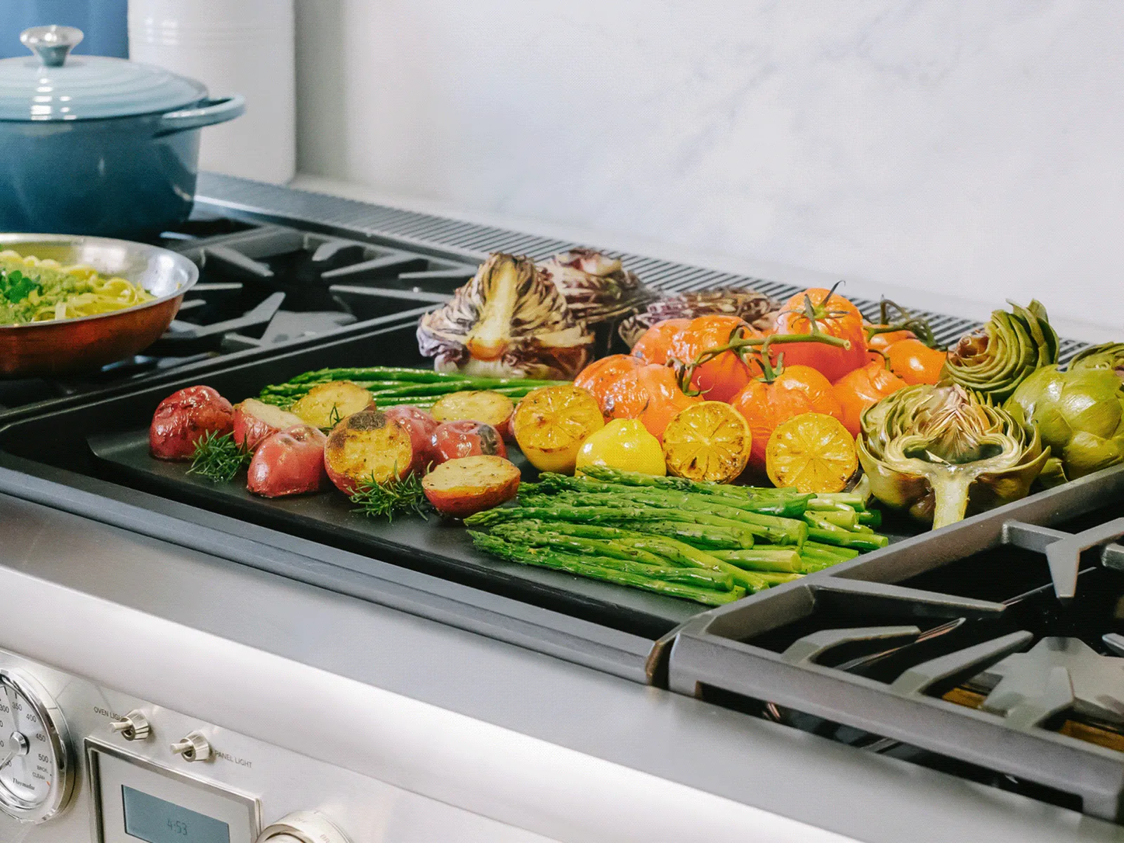 Assorted fresh vegetables including asparagus, tomatoes, and artichokes roasting on a grill pan over a stovetop.