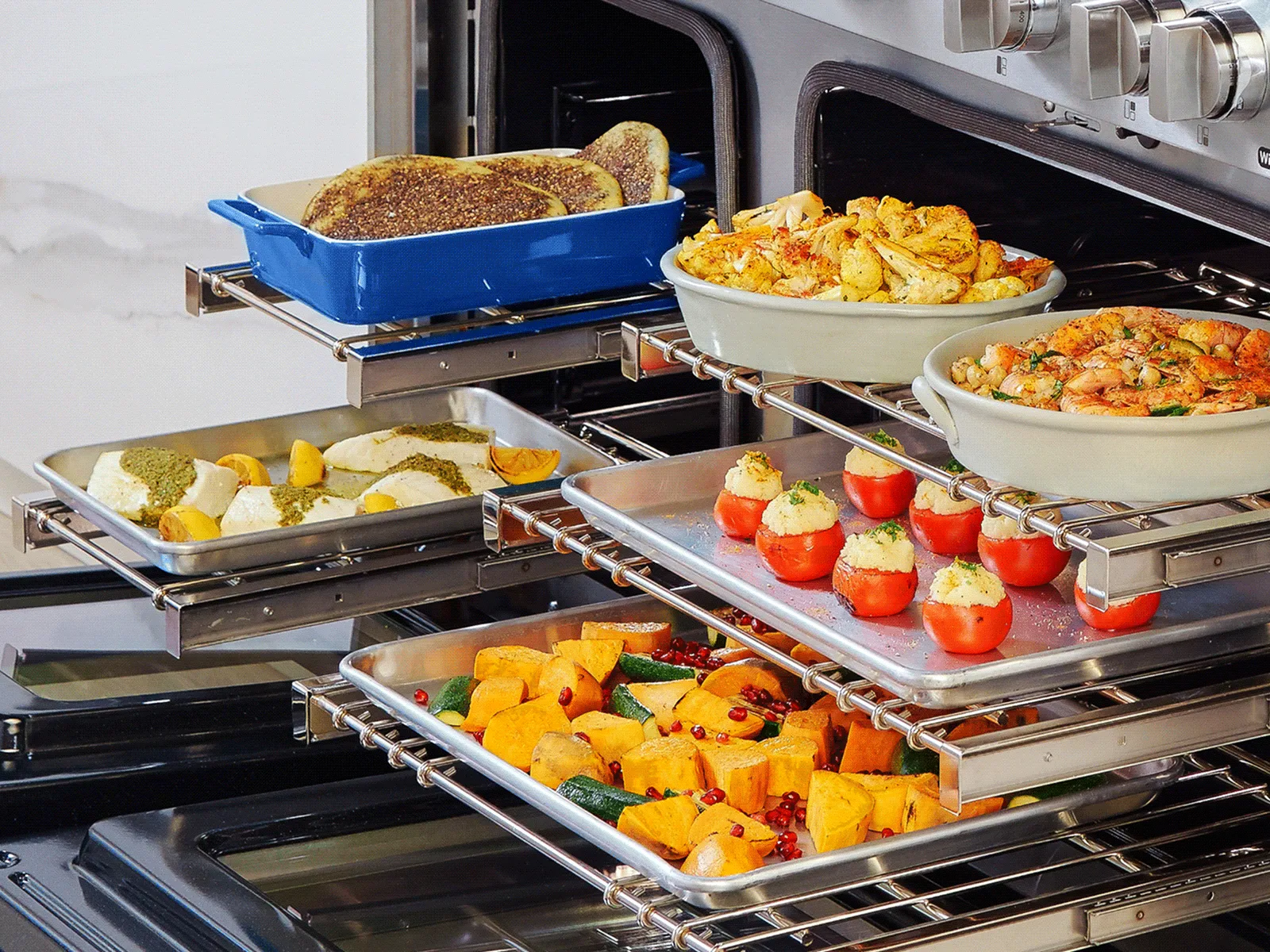 Oven open with multiple racks holding trays of cooked vegetables, stuffed tomatoes, shrimp, and bread.