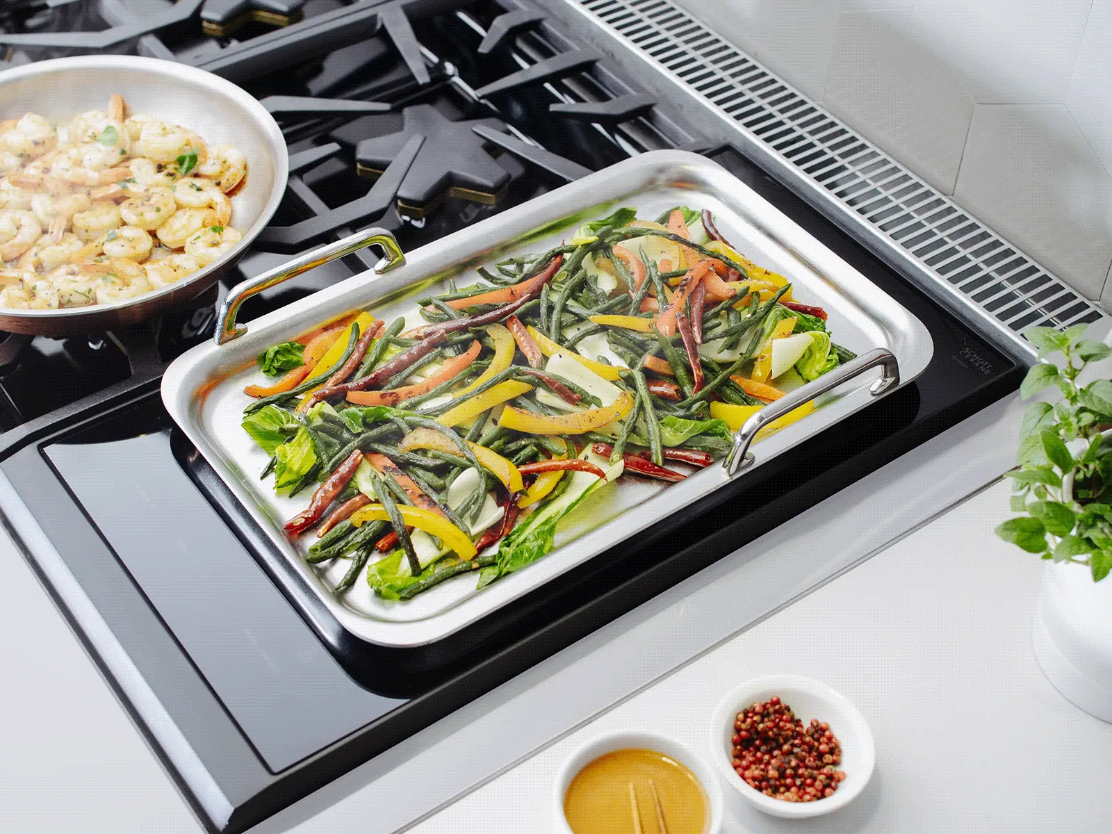 Tray of roasted colorful vegetables (zucchini, red and yellow peppers, onions) on a stovetop, with a pan of sautéed shrimp, small bowls of sauce and peppercorns, and a potted herb nearby.