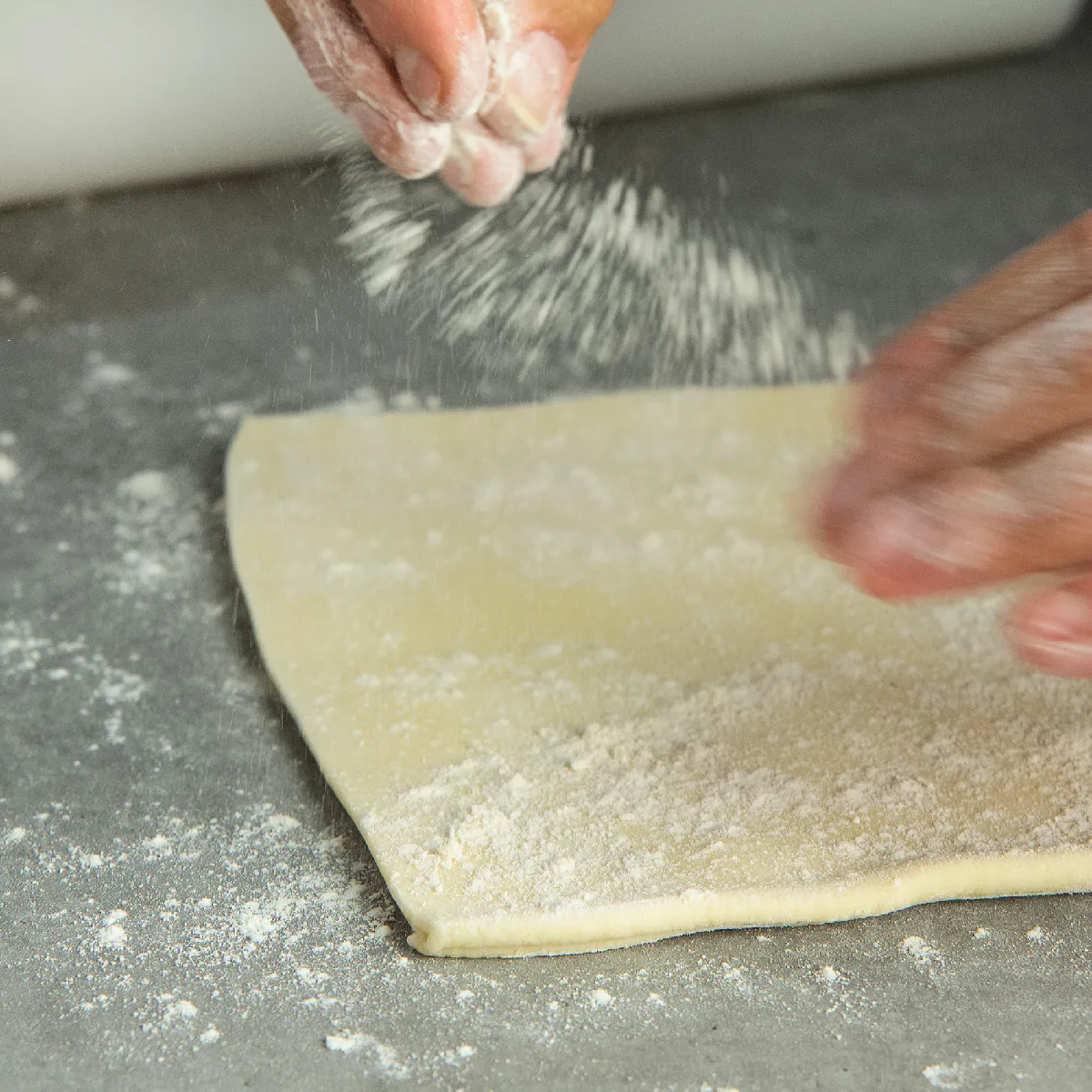 Mani che spolverano farina su pasta stesa su un piano infarinato.