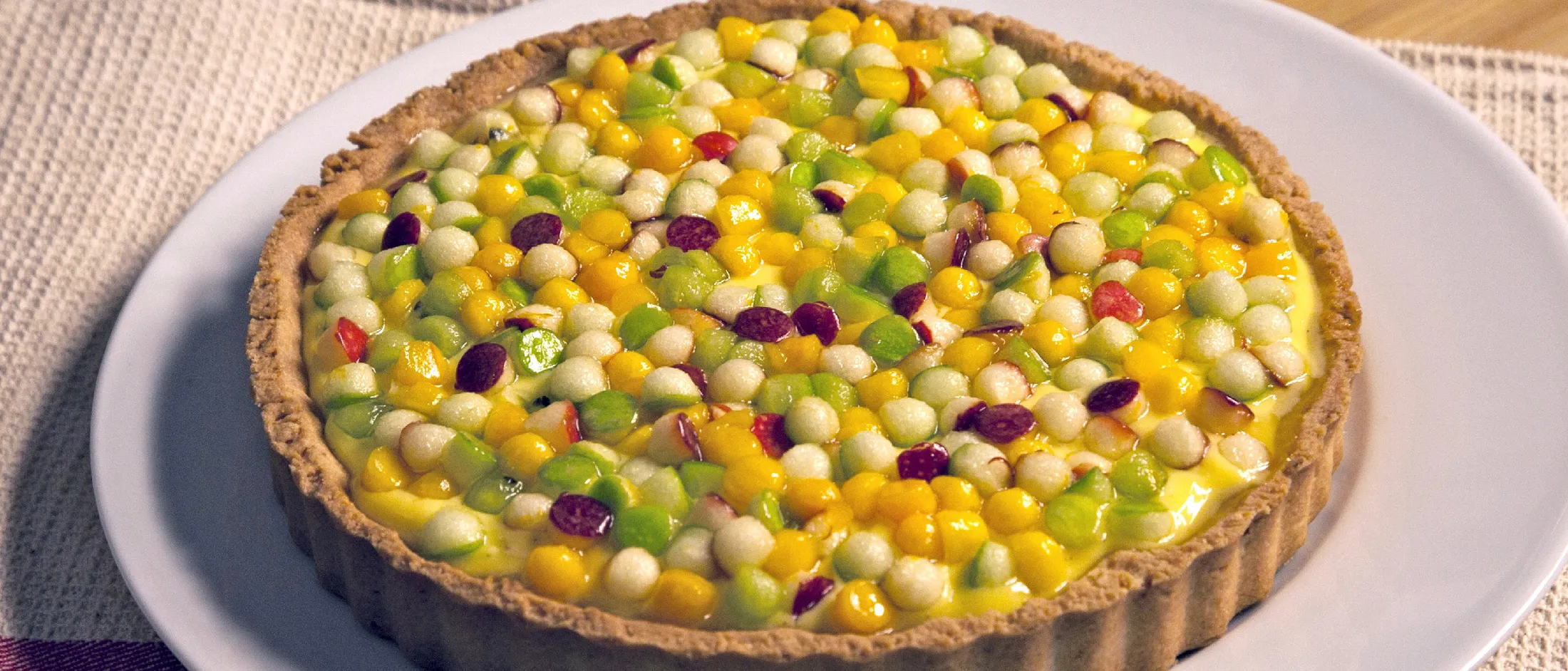 Crostata di frutta con una crosta dorata sormontata da pezzi di frutta verde, gialla, bianca e rossa, servita su un piatto bianco su una tovaglietta intrecciata