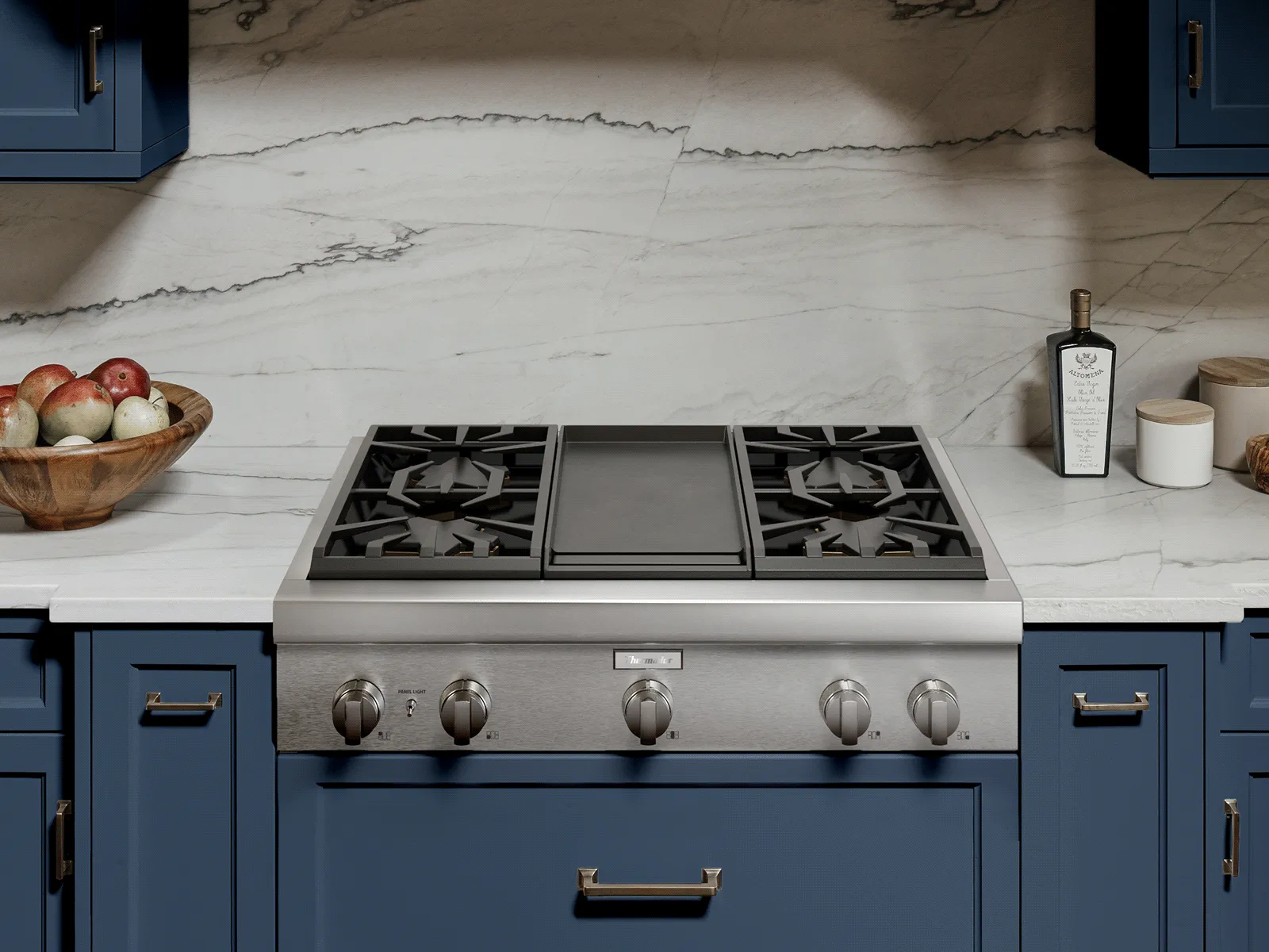 Front view of a stainless-steel gas range with a central griddle and four burners set into a white marble countertop and backsplash, flanked by blue cabinets; a wooden bowl of fruit sits on the left and an oil bottle and small container on the right.