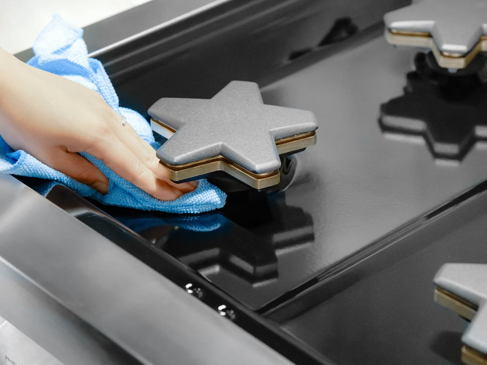 Hand wiping a star-shaped gas burner cap on a glossy black stovetop with a blue cloth.