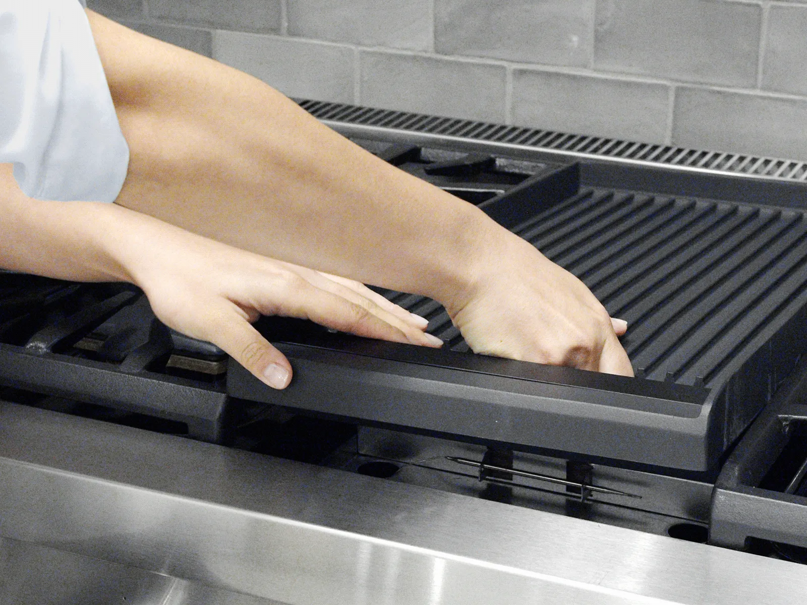 Two hands lifting a ridged cast-iron griddle from the top of a stainless-steel gas range with a tiled backsplash.