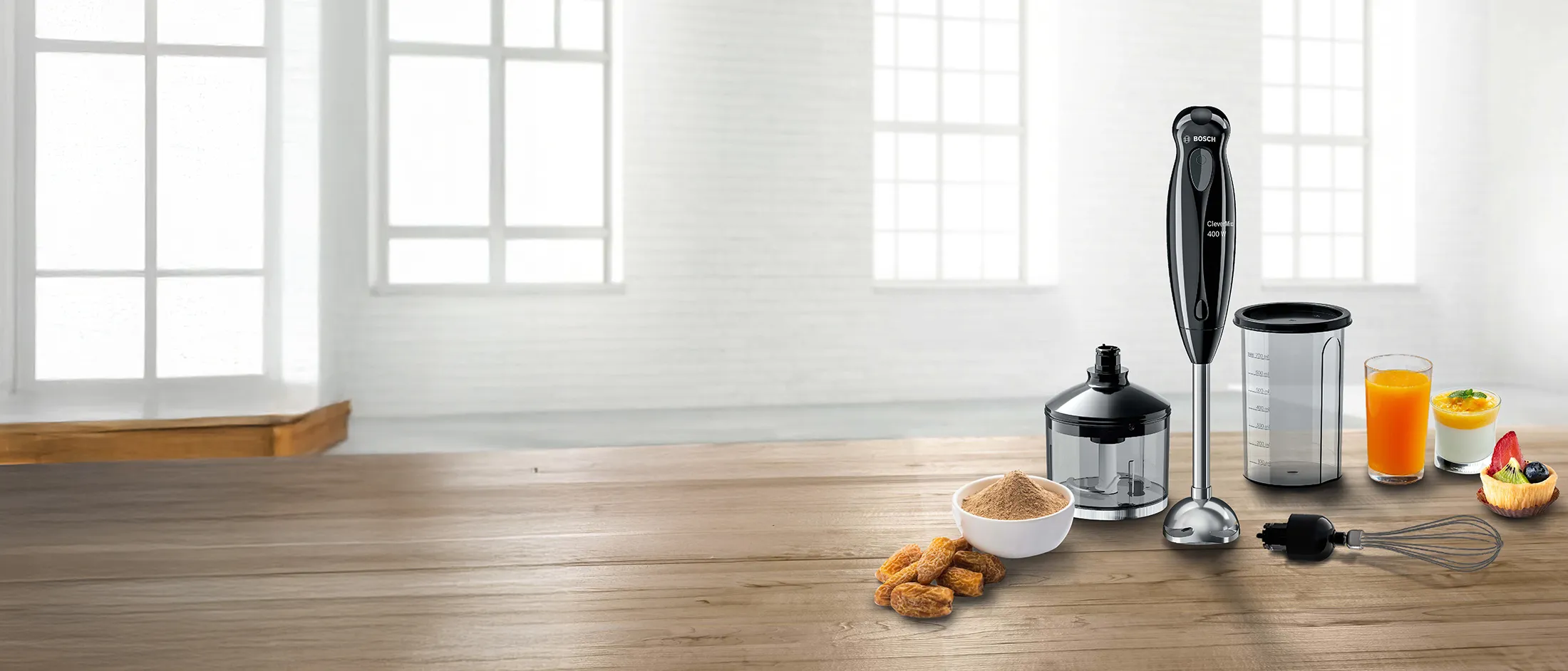 A Bosch hand blender and its accessories, including a chopper, measuring cup, whisk, and food items like cookies, juice, and a tart, on a wooden table.