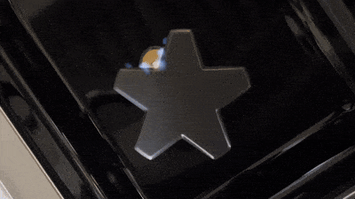 Top-down close-up of a star-shaped gas burner cap surrounded by low blue flames on a glossy black stovetop.