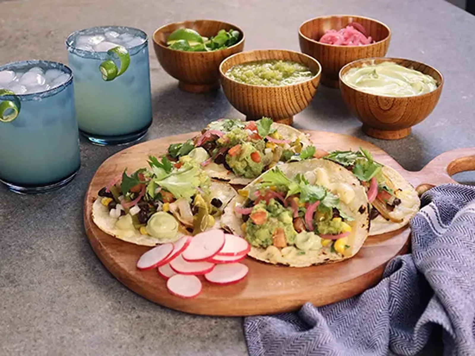 Three street tacos with various toppings, surrounded by bowls of salsas and two margaritas.