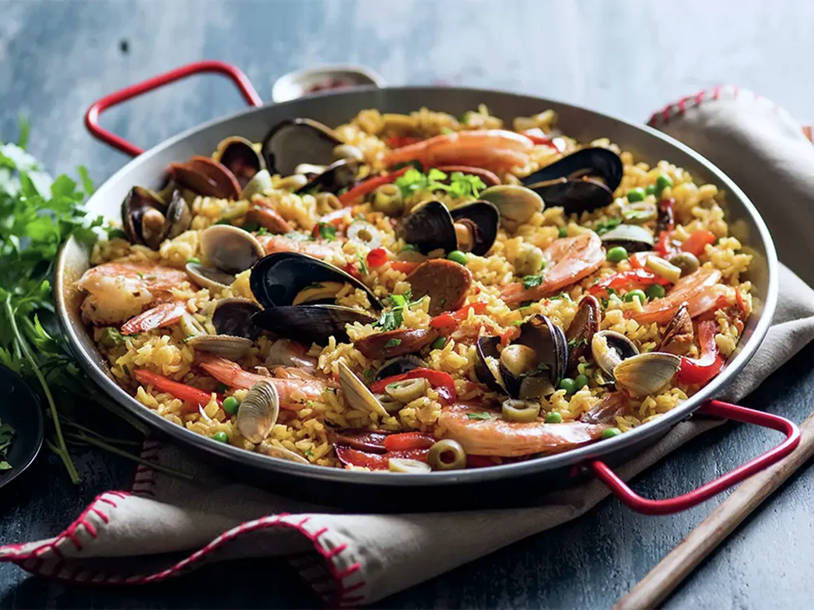 Pan of seafood paella with shrimp, mussels, clams, red peppers, olives, and rice, served on a cloth with a spoon beside it.