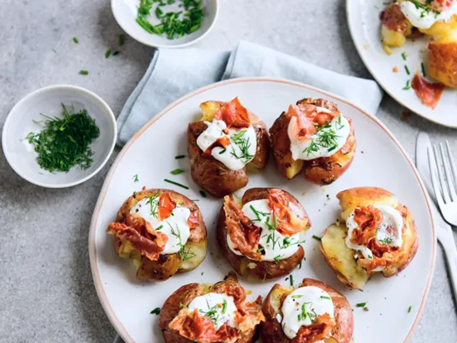 Plate of twice-baked potatoes topped with sour cream, bacon, and chopped chives, with small bowls of herbs on the side.