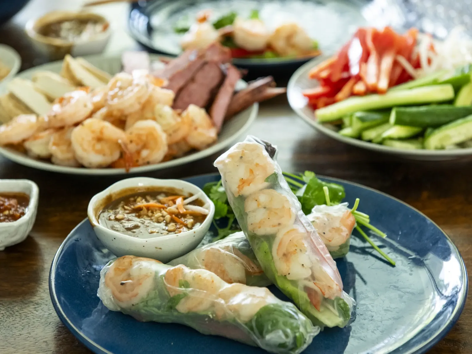 A close-up shot of a Vietnamese-style feast, centered on two fresh spring rolls filled with shrimp, greens, and vermicelli, served on a blue plate with a dipping sauce.
