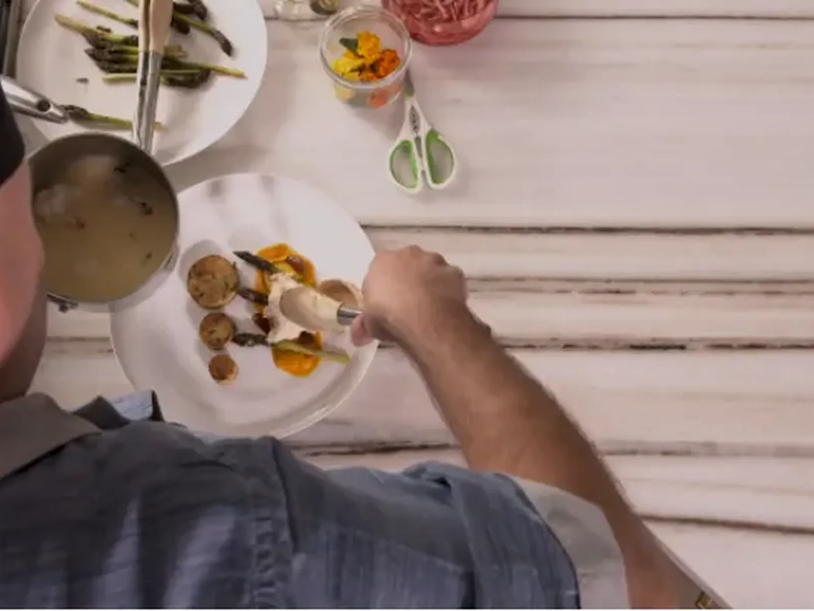 A man in a blue shirt is plating food, using tongs to arrange scallops and asparagus on a white plate.