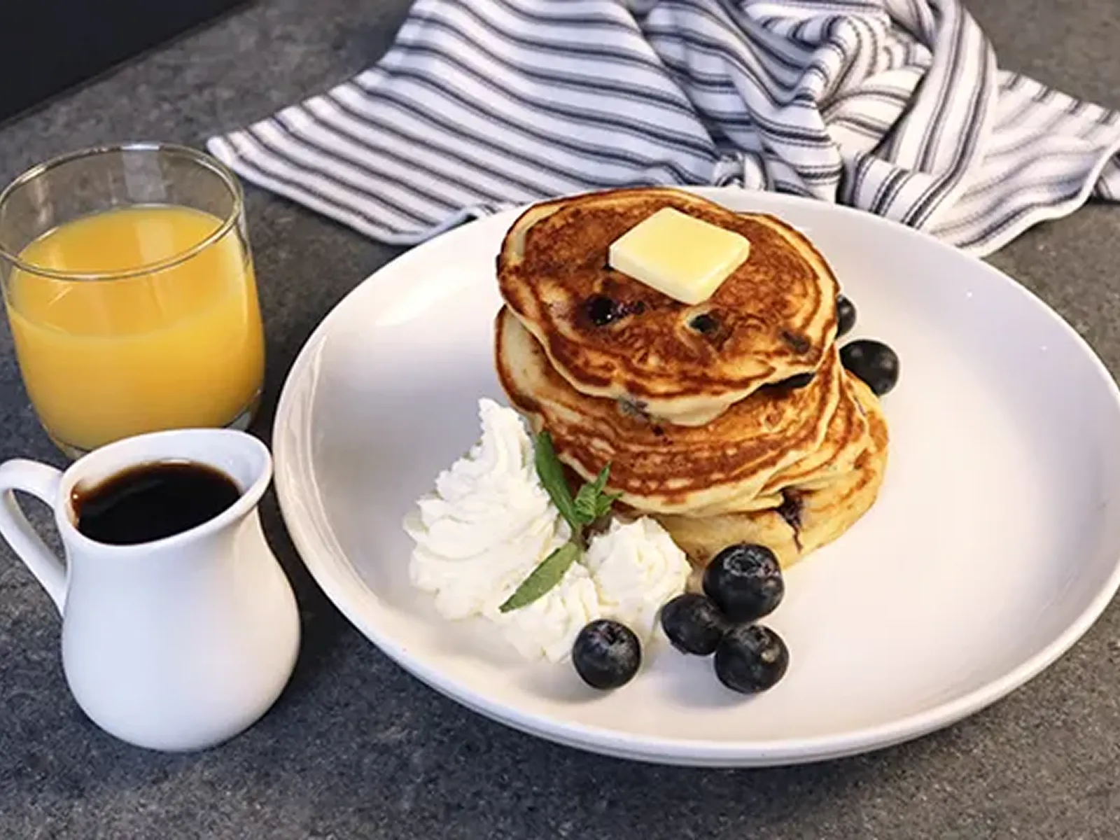 Stack of blueberry pancakes with butter, whipped cream, blueberries, orange juice, and syrup.