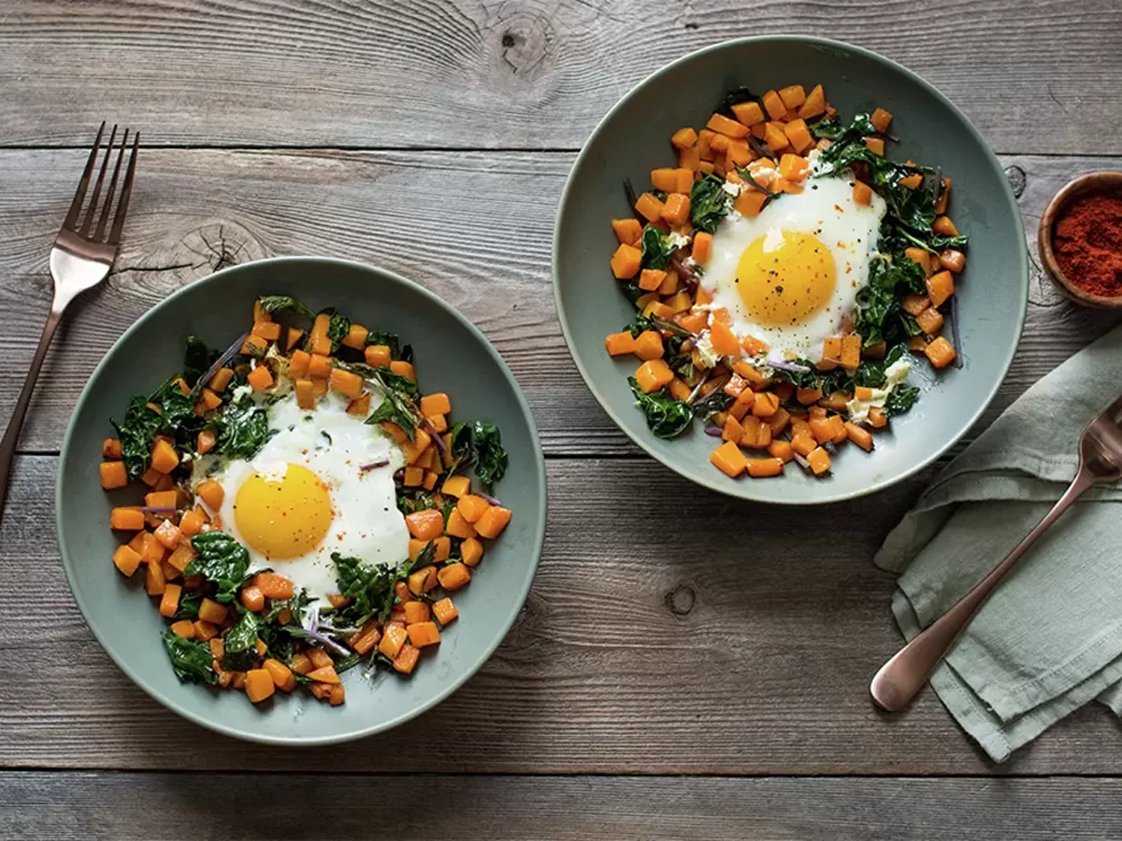 Two bowls of breakfast hash with fried eggs, sweet potatoes, and kale on a rustic wooden table.