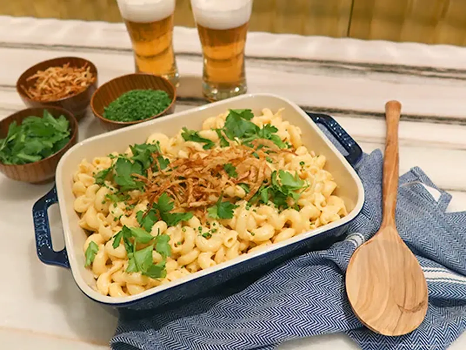 Baked macaroni and cheese topped with crispy onions and fresh herbs, served with small bowls of garnishes and two glasses of beer.