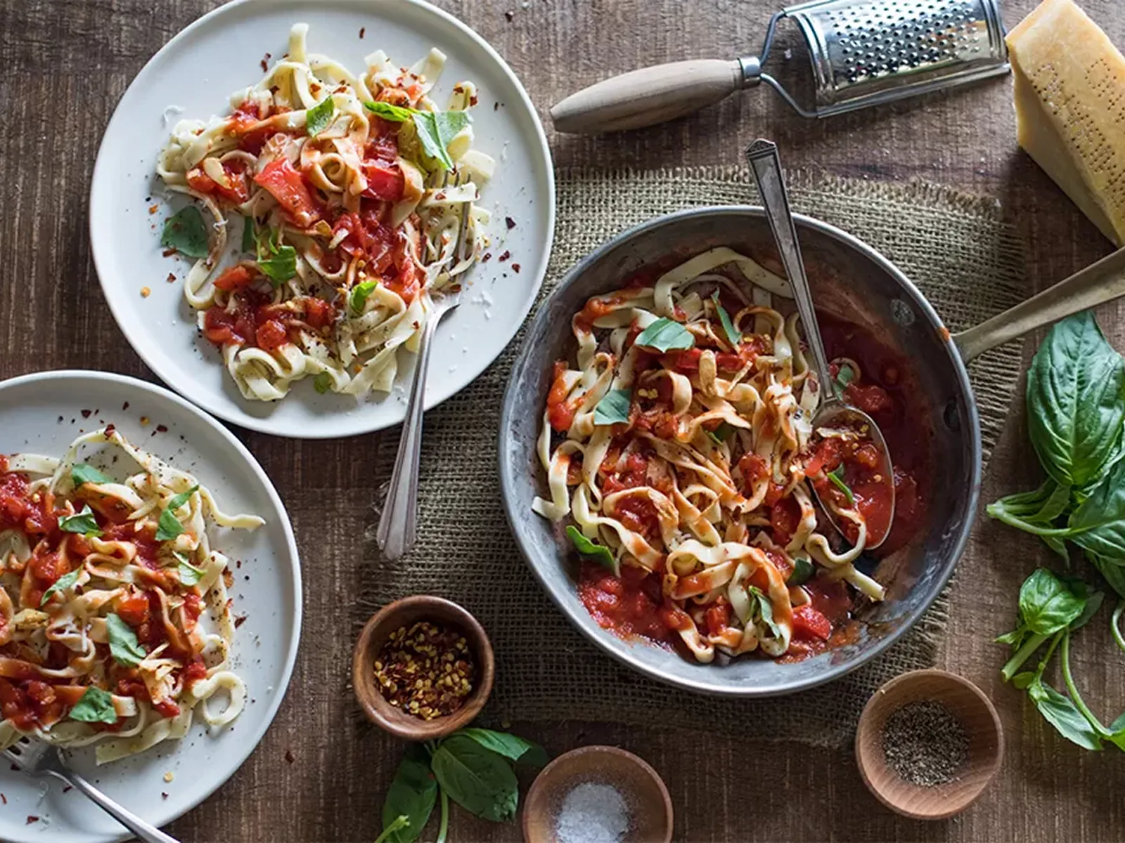 Three plates and one bowl of pasta with tomato sauce, garnished with fresh basil and red pepper flakes on a wooden table.