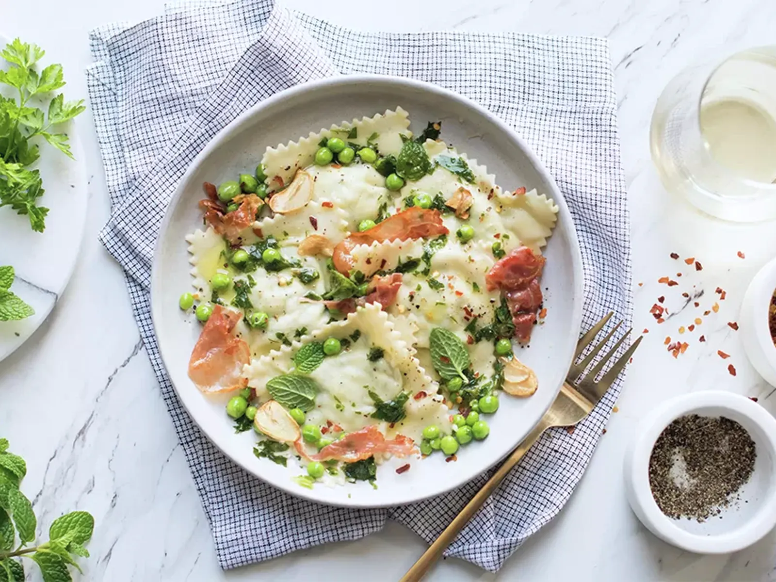 Plate of ravioli pasta with peas, crispy pancetta, garlic slices, and herbs on a gray napkin, accompanied by seasoning bowls and a glass.