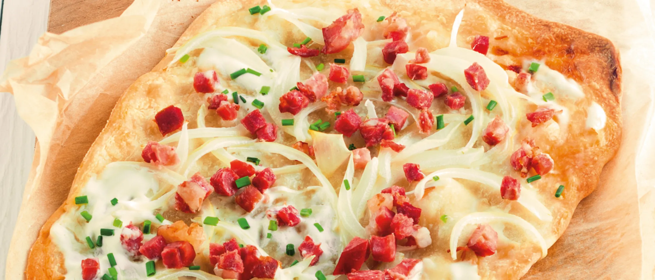 Flammkuchen auf Backpapier, belegt mit Sauerrahm, Speckwürfeln, Zwiebelringen und gehacktem Schnittlauch.