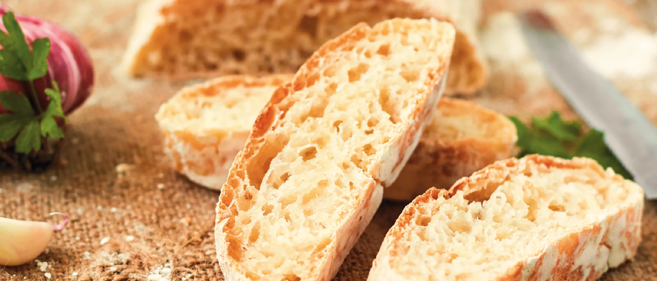 Nahaufnahme von geschnittenem, knusprigem Brot auf einem Holzbrett, im Hintergrund eine Knoblauchknolle und Petersilie.