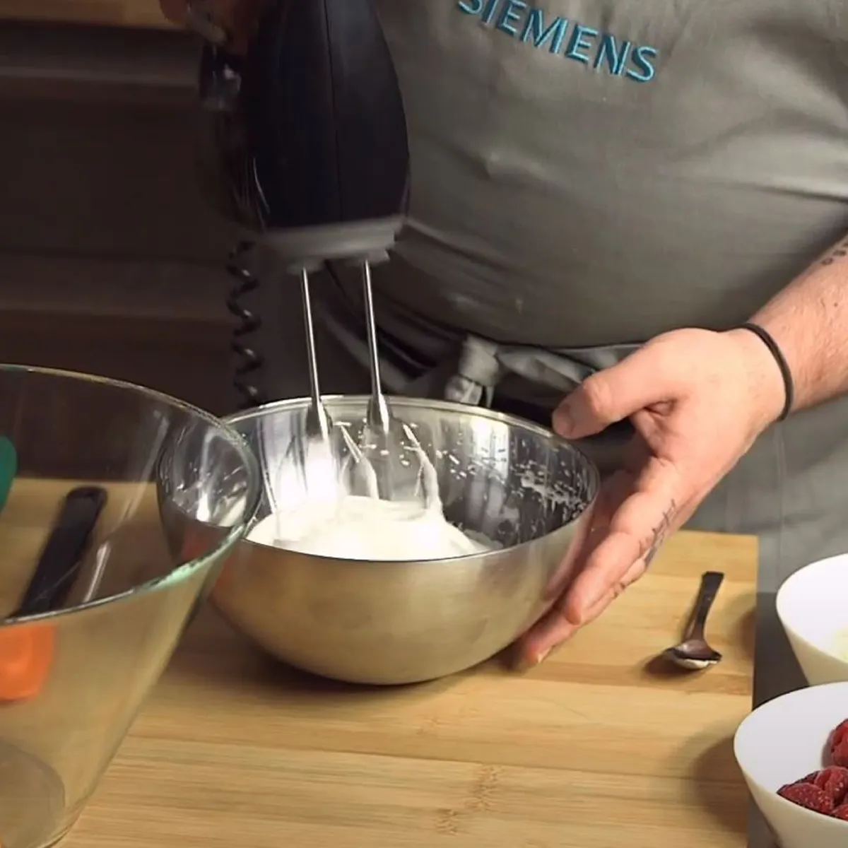 Persona con grembiule Siemens che usa un frullatore elettrico per mescolare ingredienti in una ciotola di metallo su una superficie di legno con uova e lamponi vicini