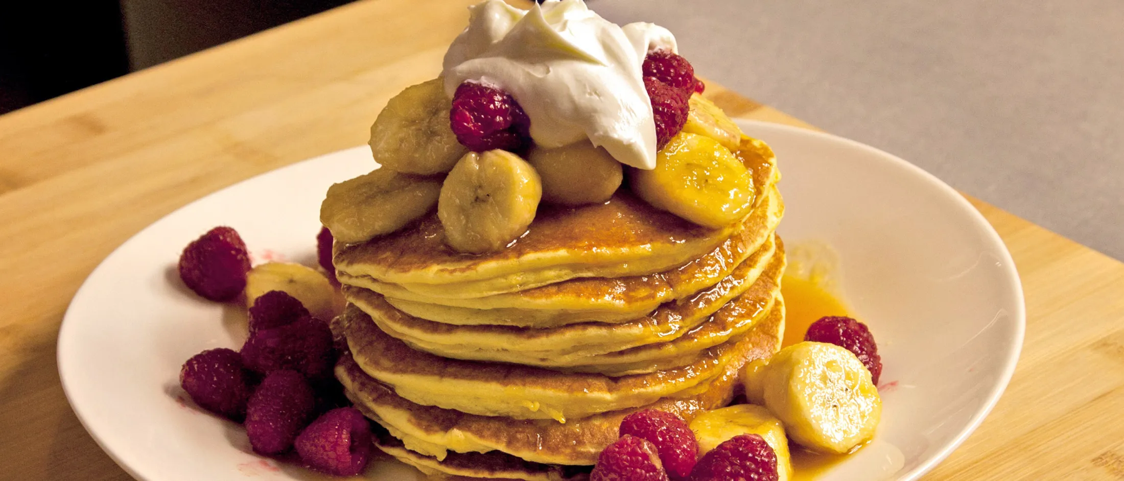Una pila di soffici pancake guarniti con fette di banana, lamponi, panna montata e sciroppo, serviti su un piatto bianco.