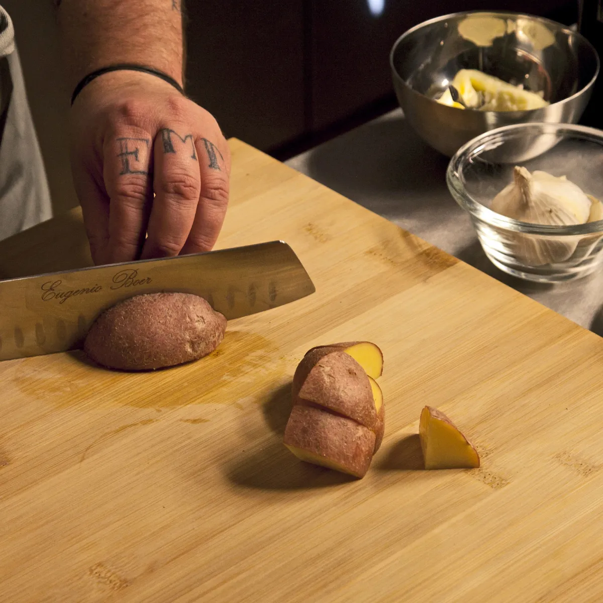 Una persona con le dita tatuate che affetta patate marroni su un tagliere di legno, con aglio e altri ingredienti accanto.