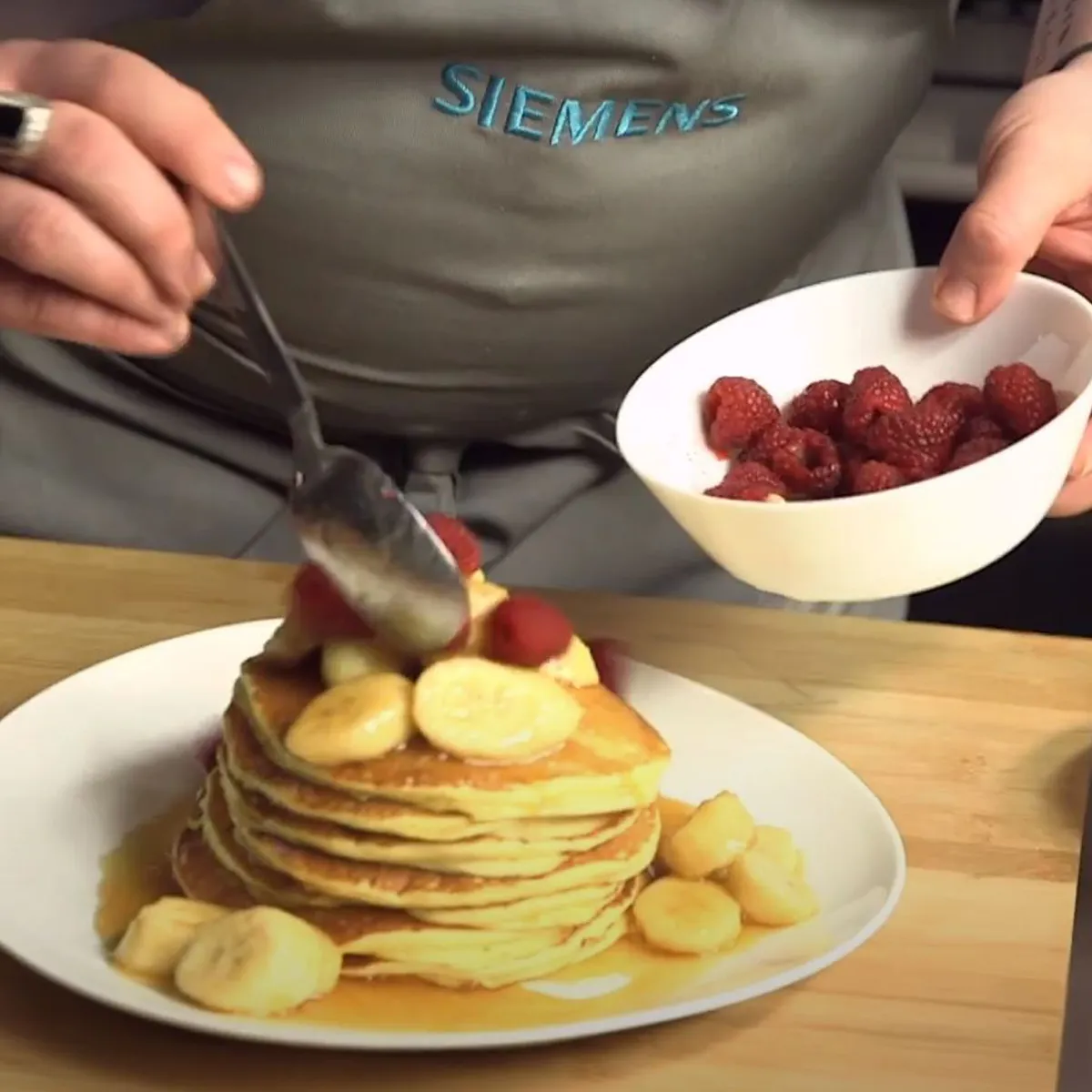 Persona che tiene una ciotola di lamponi e li versa con un cucchiaio su una pila di pancake con fette di banana su un piatto bianco