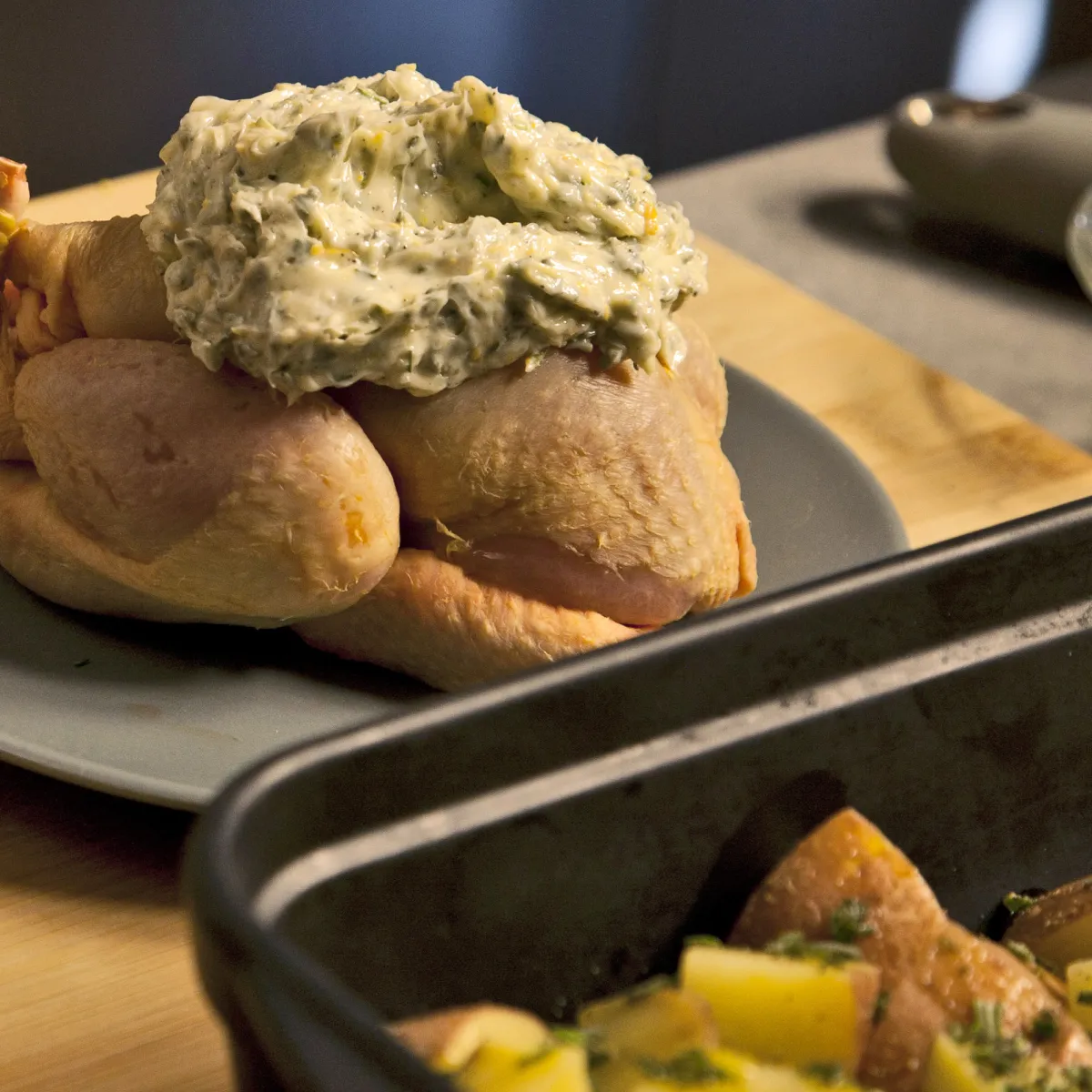 Un pollo intero crudo su un piatto, condito con burro alle erbe, con patate aromatizzate in una teglia da forno accanto, su un tagliere di legno.