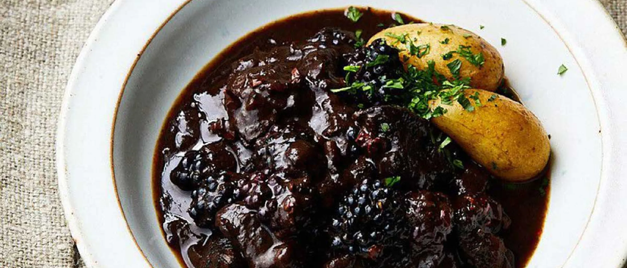 Hirschgulasch mit Kakao-Nibs und Brombeeren serviert mit Kartoffeln auf weißem Teller.