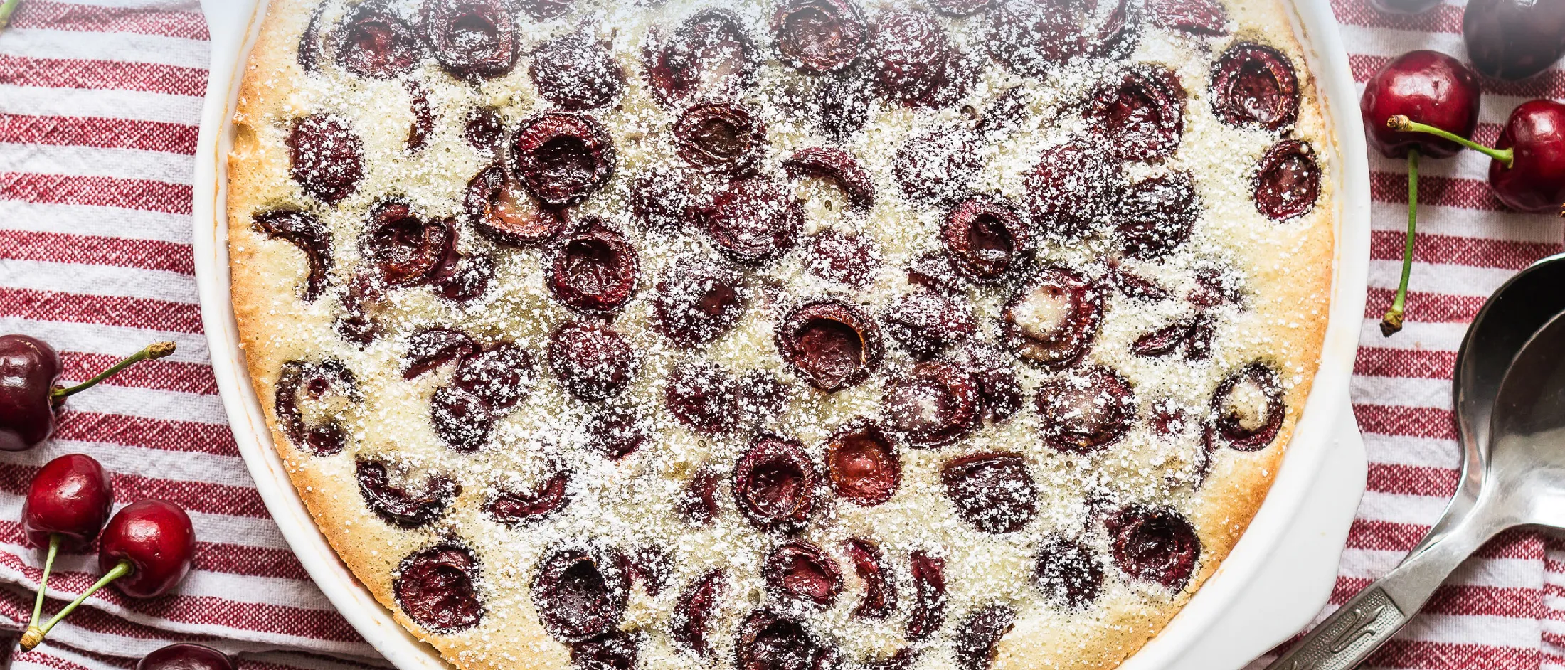 Kirsch-Grieß Kuchen auf einer weißen Platte angerichtet auf einem rot-weiß gestreiften Tuch mit Kirschen.