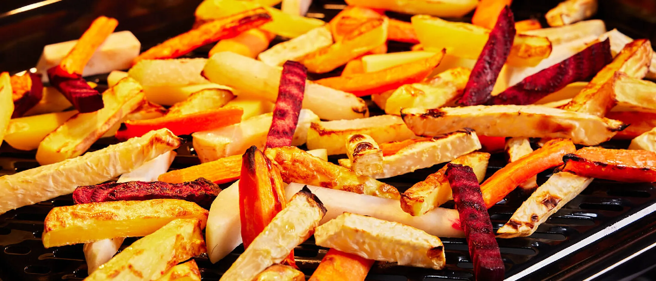 Geröstete, bunte Gemüsestäbchen, darunter Karotten, Rote Bete und Pastinaken, auf einem schwarzen Grillblech.