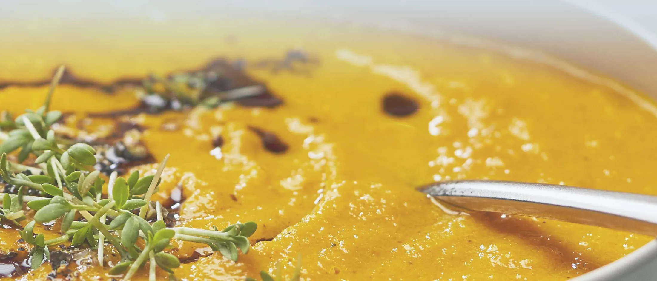 Nahaufnahme einer cremigen Kürbissuppe, garniert mit frischen Kräutern und beträufelt mit Olivenöl, ein Löffel liegt in der Schüssel.