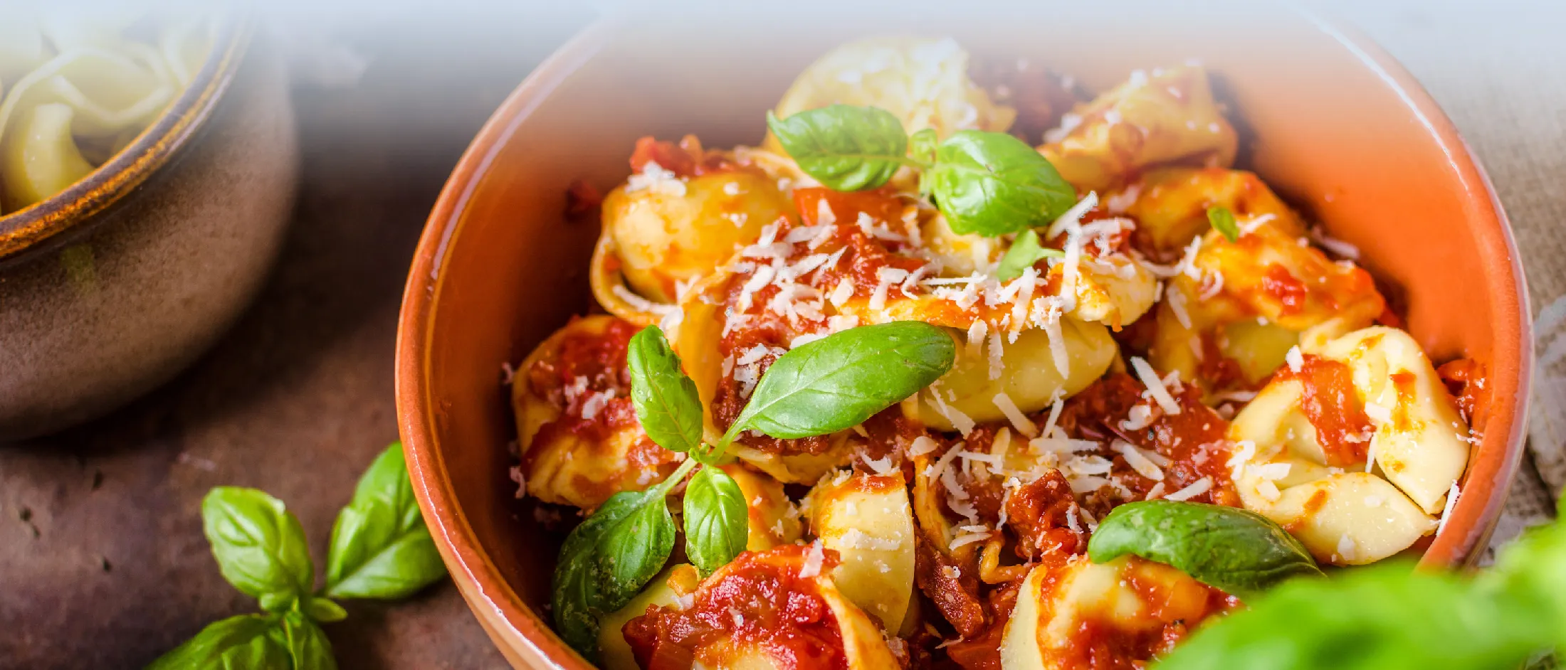 Eine Schüssel Tortellini mit Tomatensoße, geriebenem Käse und frischen Basilikumblättern auf einer rustikalen Oberfläche, daneben ein Topf mit ungekochten Nudeln.