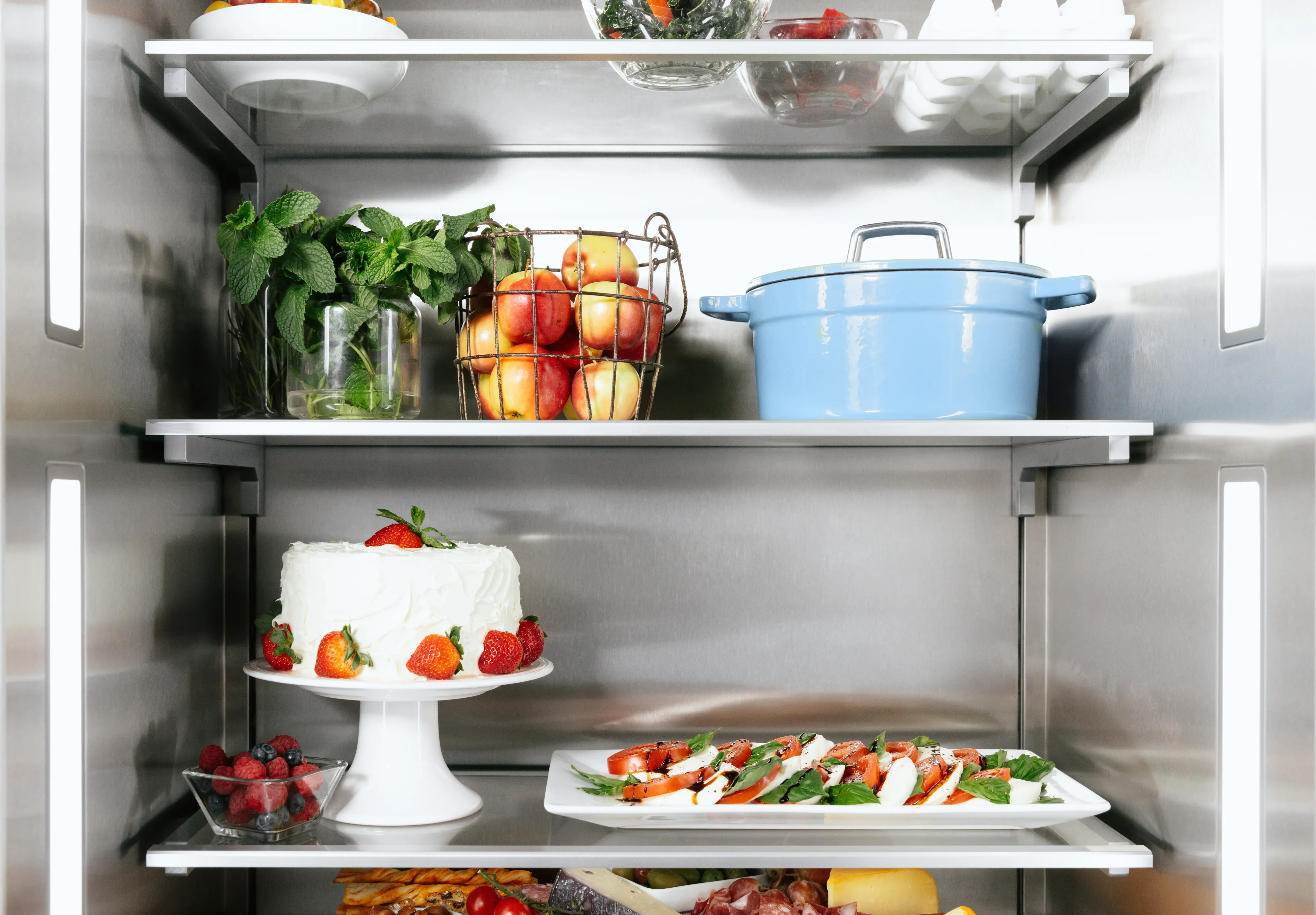Open refrigerator with a white cake, fresh strawberries, a Caprese salad, apples in a basket, a blue pot, and leafy greens.