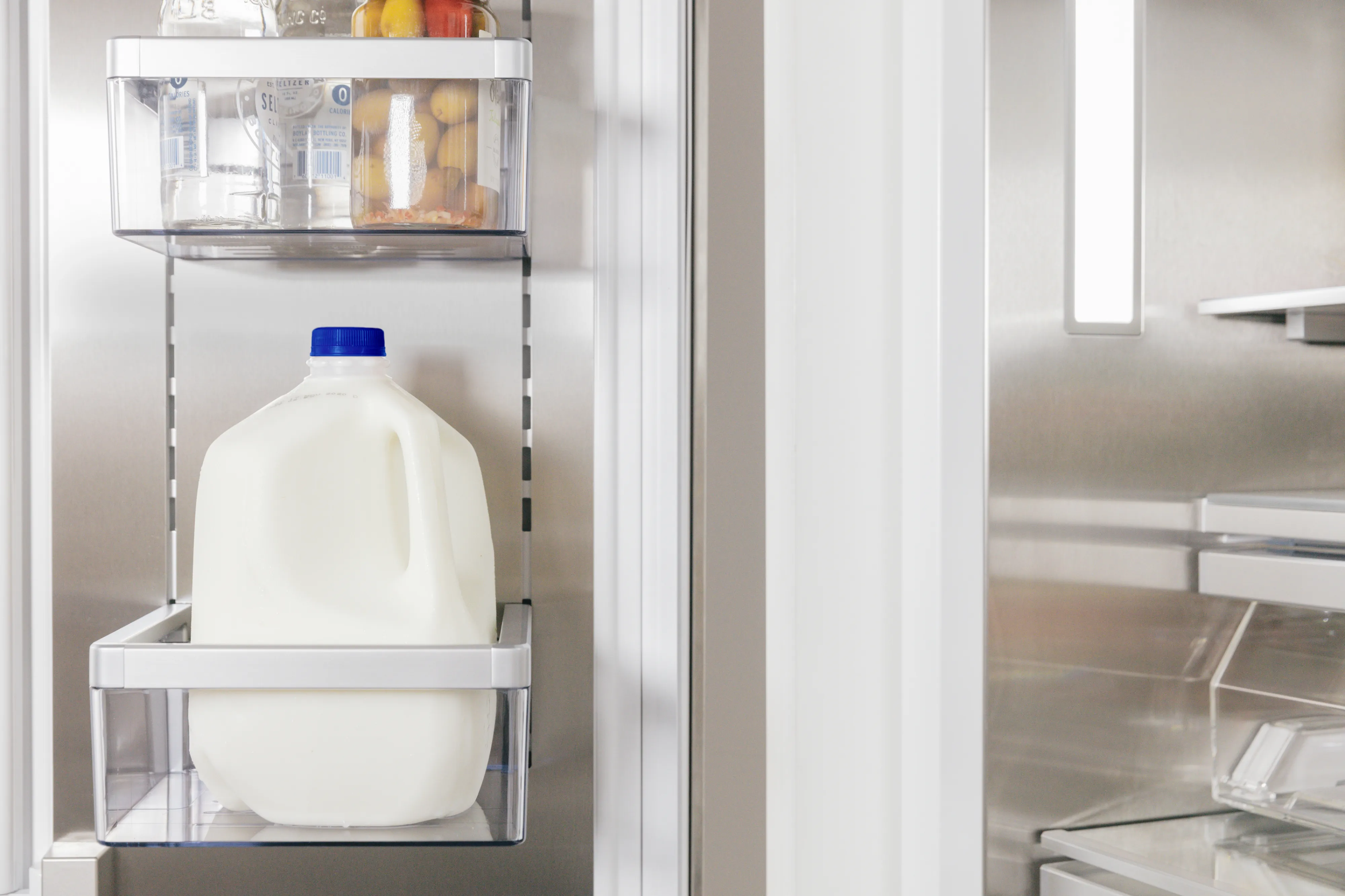 Interior of an open refrigerator door focusing on a clear plastic shelf holding a gallon jug of milk with a blue cap; a small clear bin with condiments sits on the shelf above and the stainless-steel refrigerator interior is visible to the right.