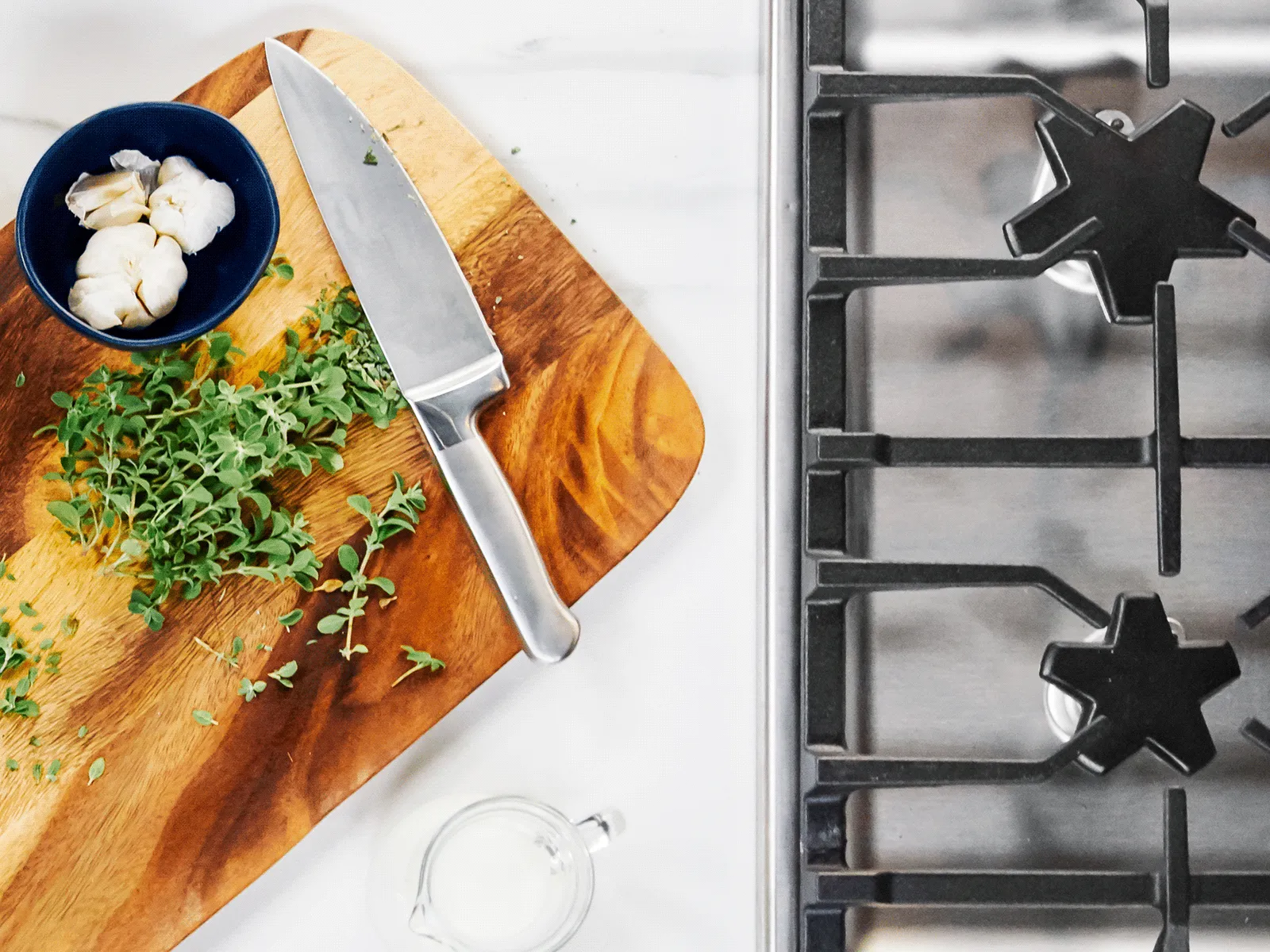 Overhead shot of a cutting board with herbs, a knife, and garlic cloves next to a stove.