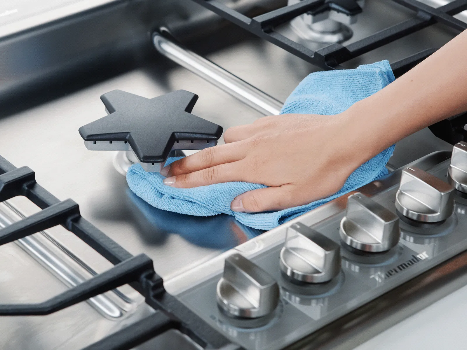 Hand cleaning a stainless steel gas stovetop with a blue cloth.