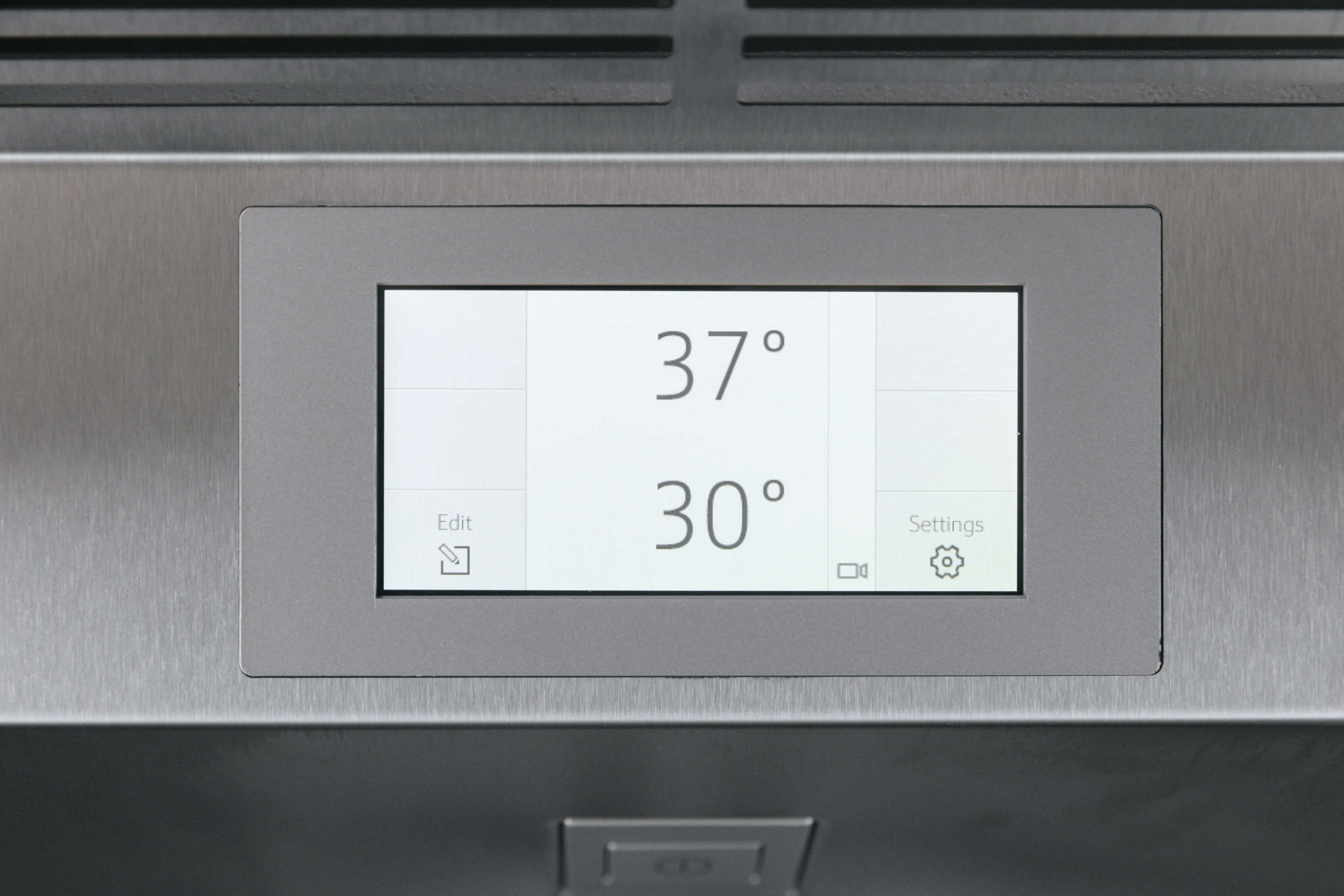 Close-up of a stainless-steel refrigerator touchscreen showing two temperatures—"37°" at the top and "30°" below—with small edit and settings icons in the lower corners.