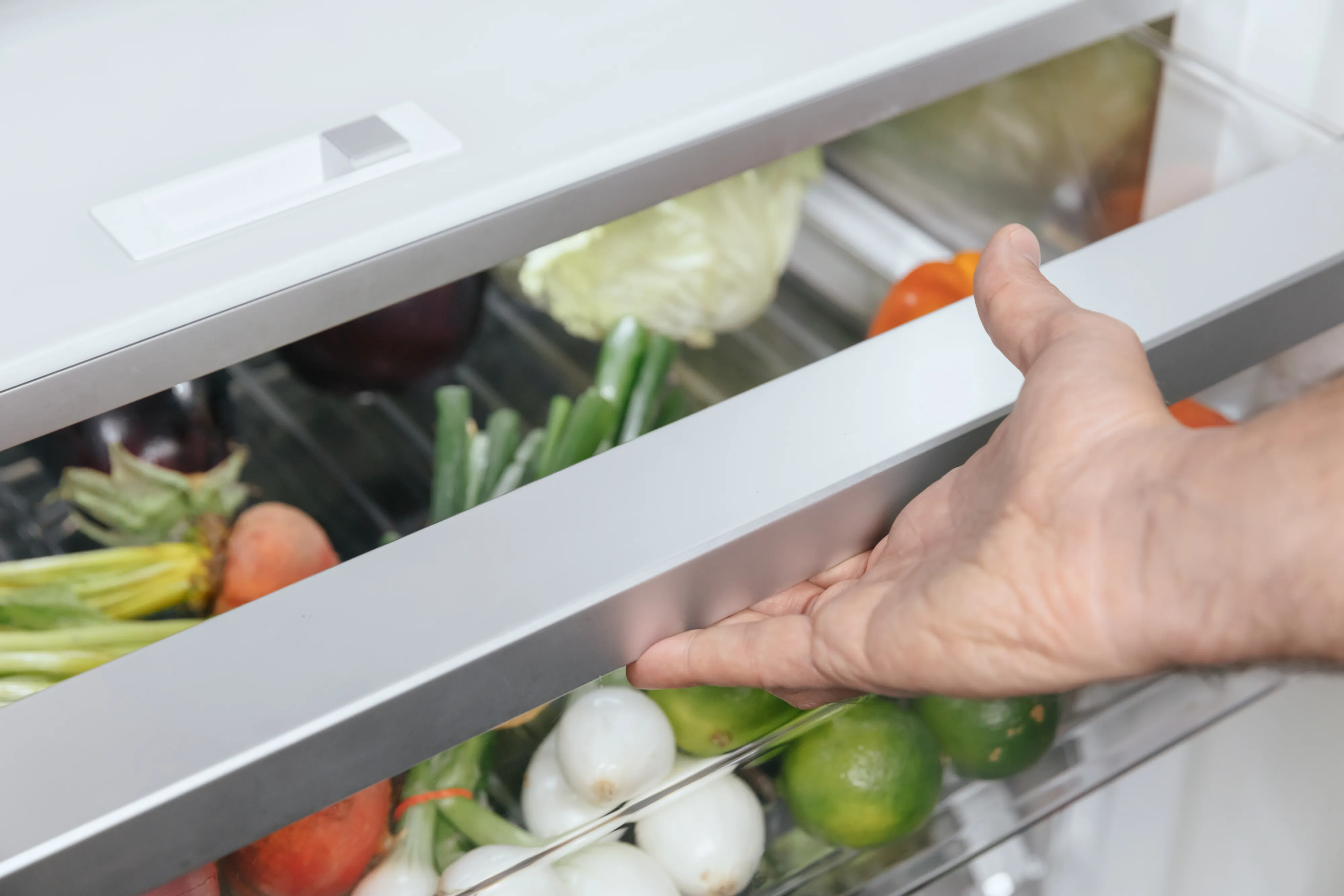 Hand pulling open a refrigerator crisper drawer filled with assorted fresh vegetables — carrots, limes, small white onions, and leafy greens visible through